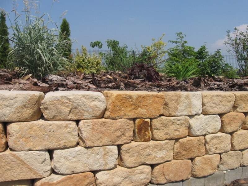Murek oporowy z piaskowca w ogrodzie, za nim roślinność i siatka ogrodzeniowa, w tle błękitne niebo.