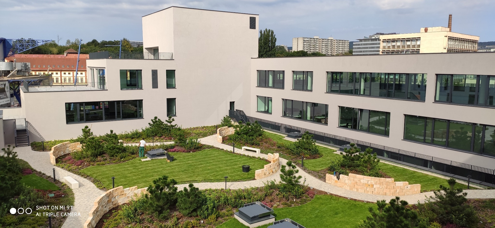 Widok z góry na zadbany, zielony dziedziniec pomiędzy nowoczesnymi budynkami biurowymi z regularnie przystrzyżonym trawnikiem, ścieżkami z jasnego kamienia i niskimi murkami z piaskowca. Pracownik...