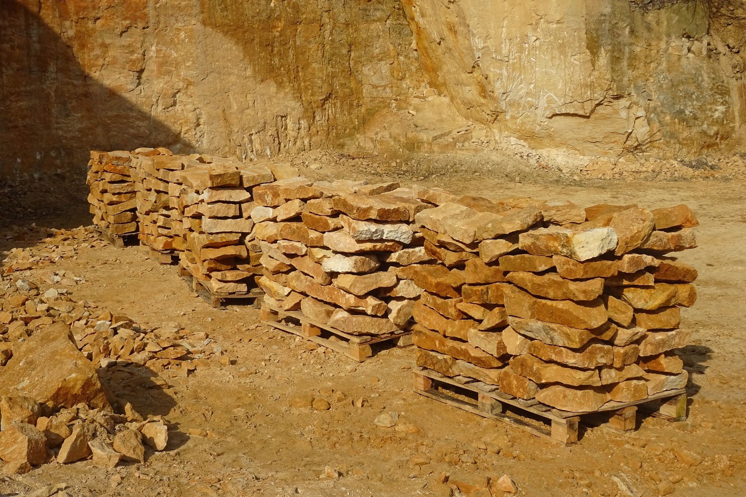 Ułożone na paletach bloki kamienne w odcieniach beżu i brązu, widoczne na tle skalnej ściany w kamieniołomie, oświetlone mocnym, naturalnym światłem.