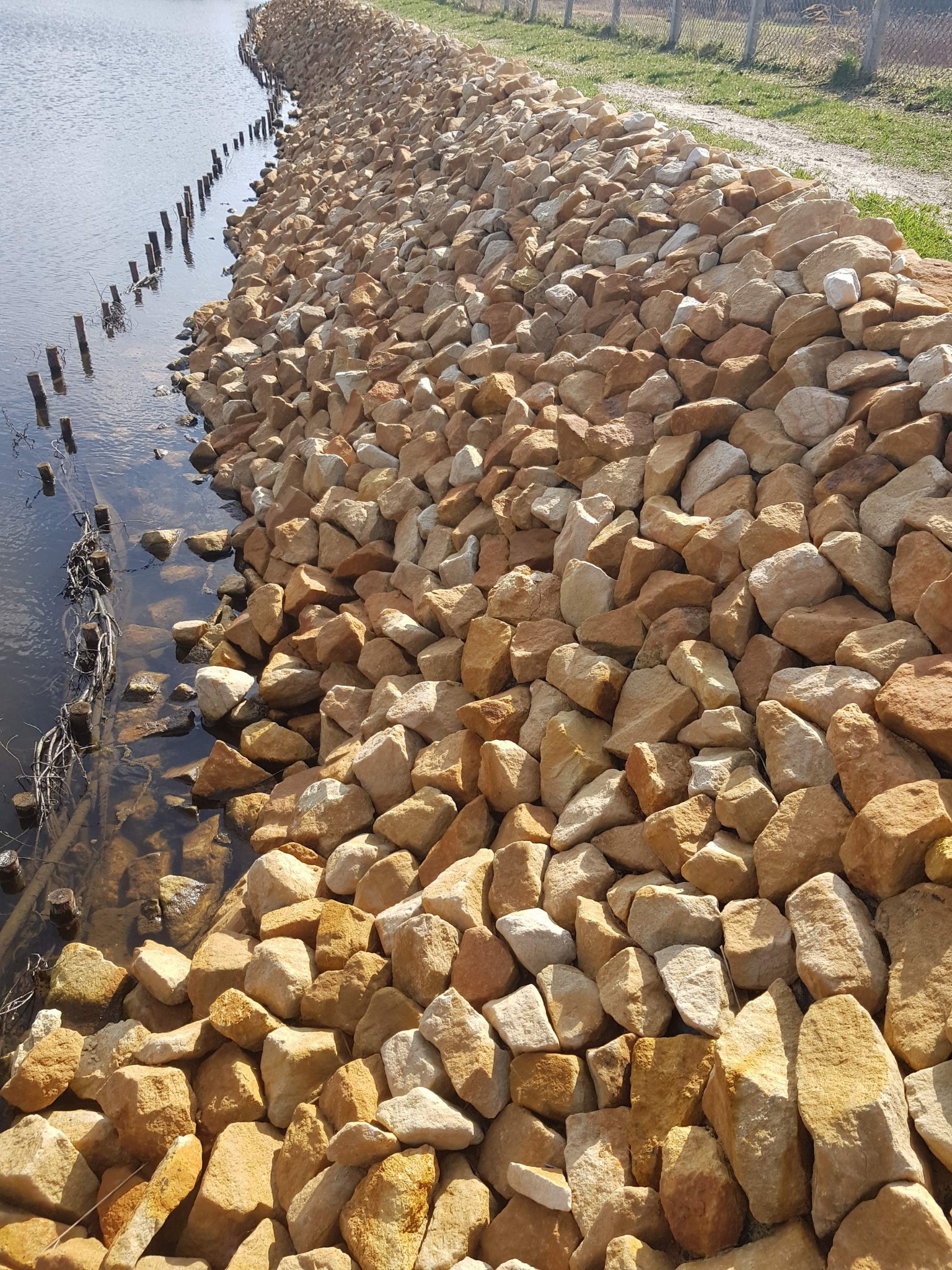 Skarpa stawu wzmocniona kamieniem łamanym hydrotechnicznym w odcieniach beżu i brązu, widoczne drewniane pale w wodzie, zarys ścieżki i ogrodzenia w tle.