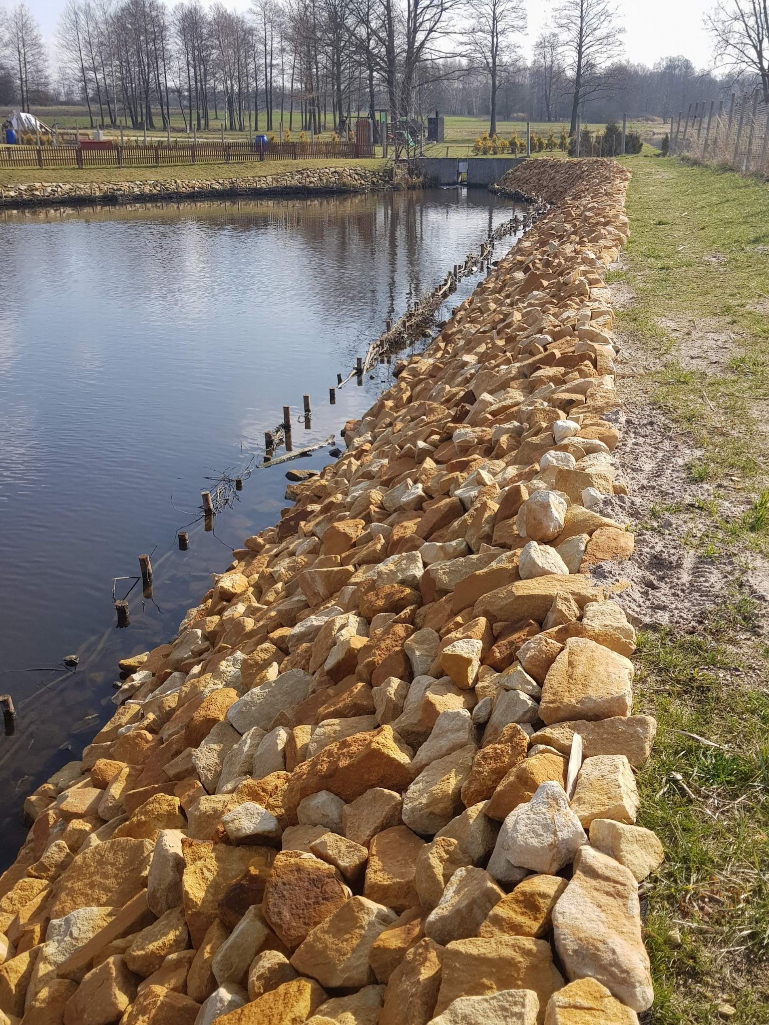 Brzeg stawu umocniony piaskowcem w odcieniach beżu i brązu, z wbitymi w dno drewnianymi palami, w tle drzewa bezlistne.