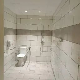 Wykończona łazienka z biało-szarymi płytkami w układzie cegiełki, wyposażona w toaletę, prysznic z deszczownicą i uchwyty dla niepełnosprawnych.