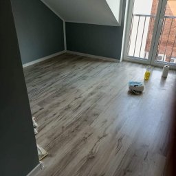 Usługi remontowo wykończeniowe Mateusz Mielcarek - Pomalowane na szaro poddasze z drewnopodobną podłogą, widoczne akcesoria malarskie i okno balkonowe z widokiem na budynek z czerwonej cegły.