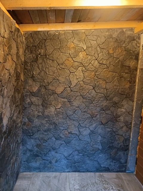 Ściana wykończona dekoracyjną okładziną imitującą kamień naturalny, z widoczną strukturą i nieregularnym kształtem elementów, w odcieniach szarości i grafitu, oświetlona od góry ciepłym światłem.