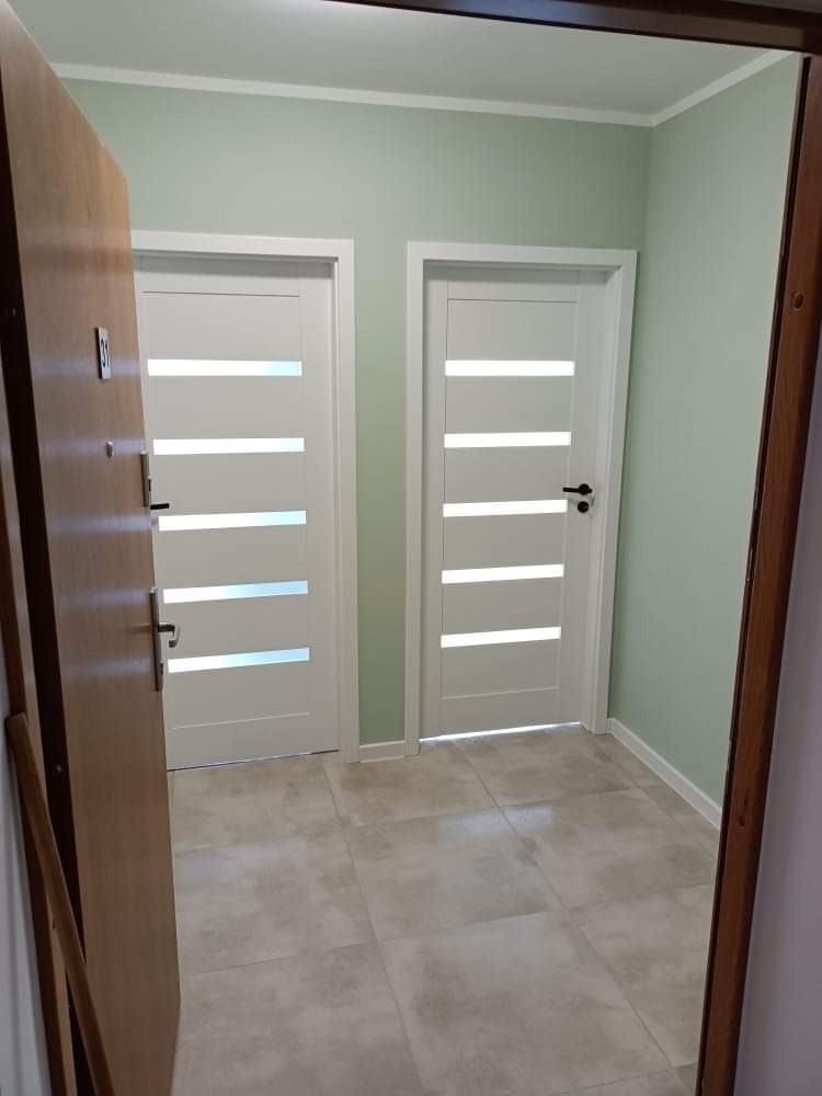 Korytarz po remoncie z dwoma białymi drzwiami z poziomymi przeszkleniami, ściany pomalowane na jasny zielony kolor, podłoga wyłożona płytkami w odcieniach szarości.