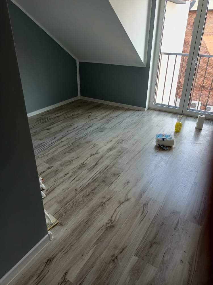 Pomalowane na szaro poddasze z drewnopodobną podłogą, widoczne akcesoria malarskie i okno balkonowe z widokiem na budynek z czerwonej cegły.