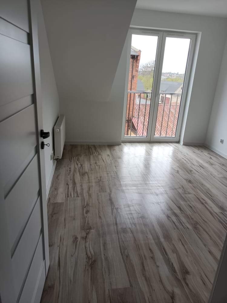 Pusty pokój na poddaszu z nową podłogą laminowaną w odcieniach szarości, świeżo pomalowanymi białymi ścianami, grzejnikiem i białymi drzwiami balkonowymi z widokiem na budynki i drzewa.