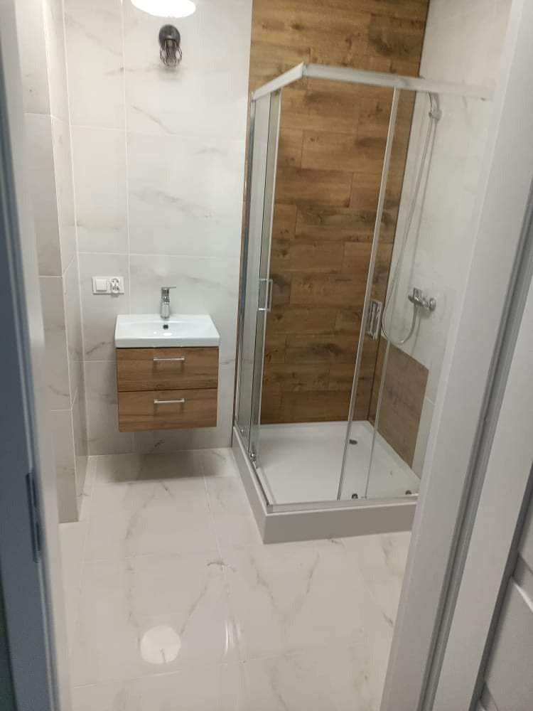 Nowoczesna łazienka z kabiną prysznicową z przezroczystego szkła, drewnopodobną ścianą w kabinie, umywalką z szafką w odcieniach drewna i marmurowymi płytkami na podłodze i ścianach.