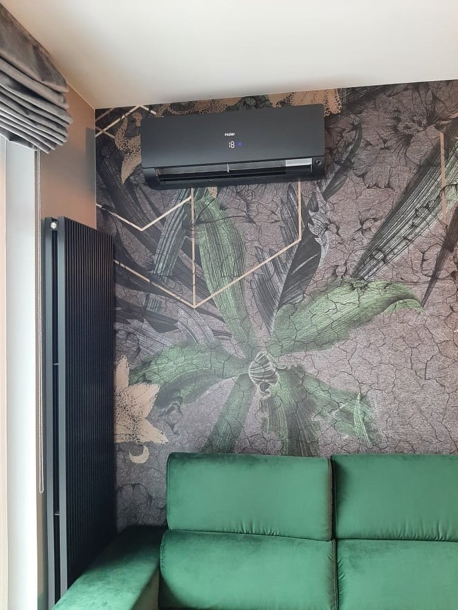 Czarny klimatyzator Haier zamontowany na ścianie z wzorem roślinnym, obok pionowego grzejnika i zielonej sofy.