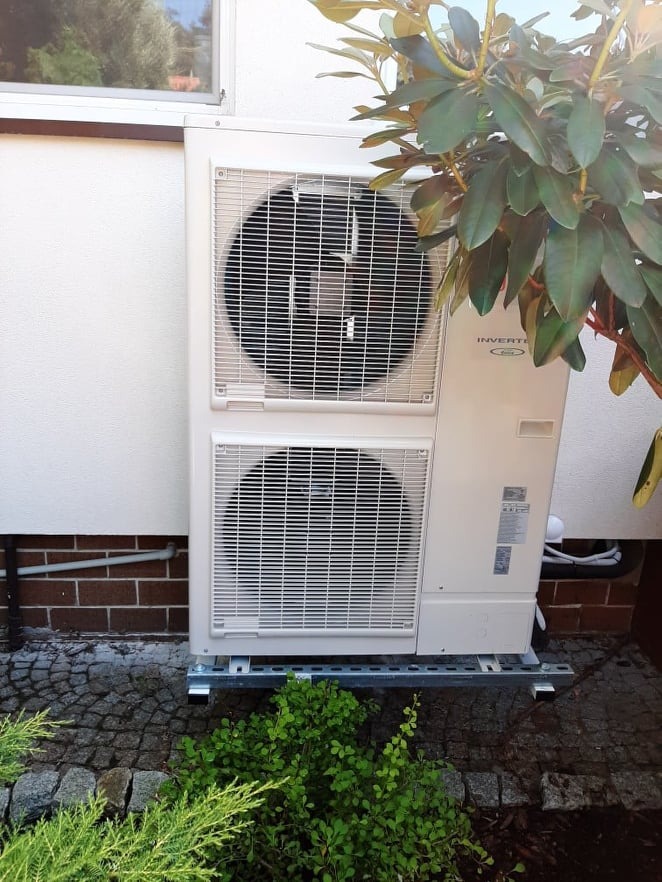 Zewnętrzna jednostka klimatyzacji typu split, zamontowana na metalowej konstrukcji przy ścianie budynku z ceglanym cokołem, otoczona zielenią.