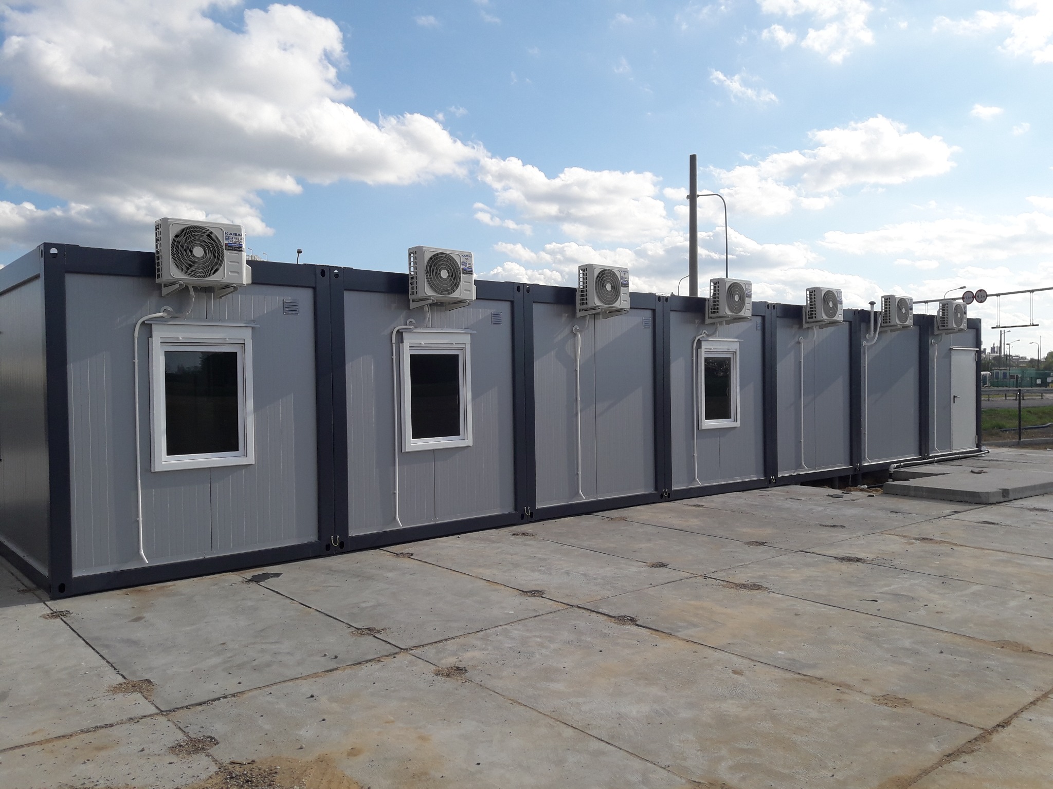 Szereg szarych kontenerów biurowych z zamontowanymi klimatyzatorami na dachu, widoczne białe ramy okien i betonowa nawierzchnia przed nimi.