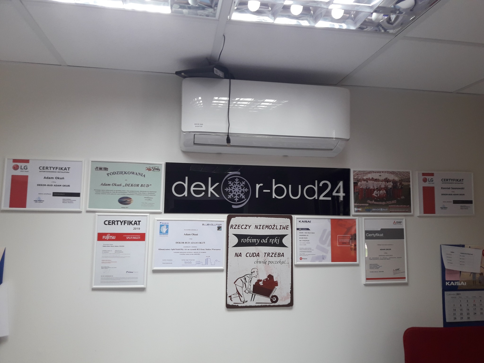 Biała klimatyzacja ścienna w biurze, otoczona certyfikatami i podziękowaniami firmy Dekr-bud24, widoczne logo firmy i humorystyczny plakat.