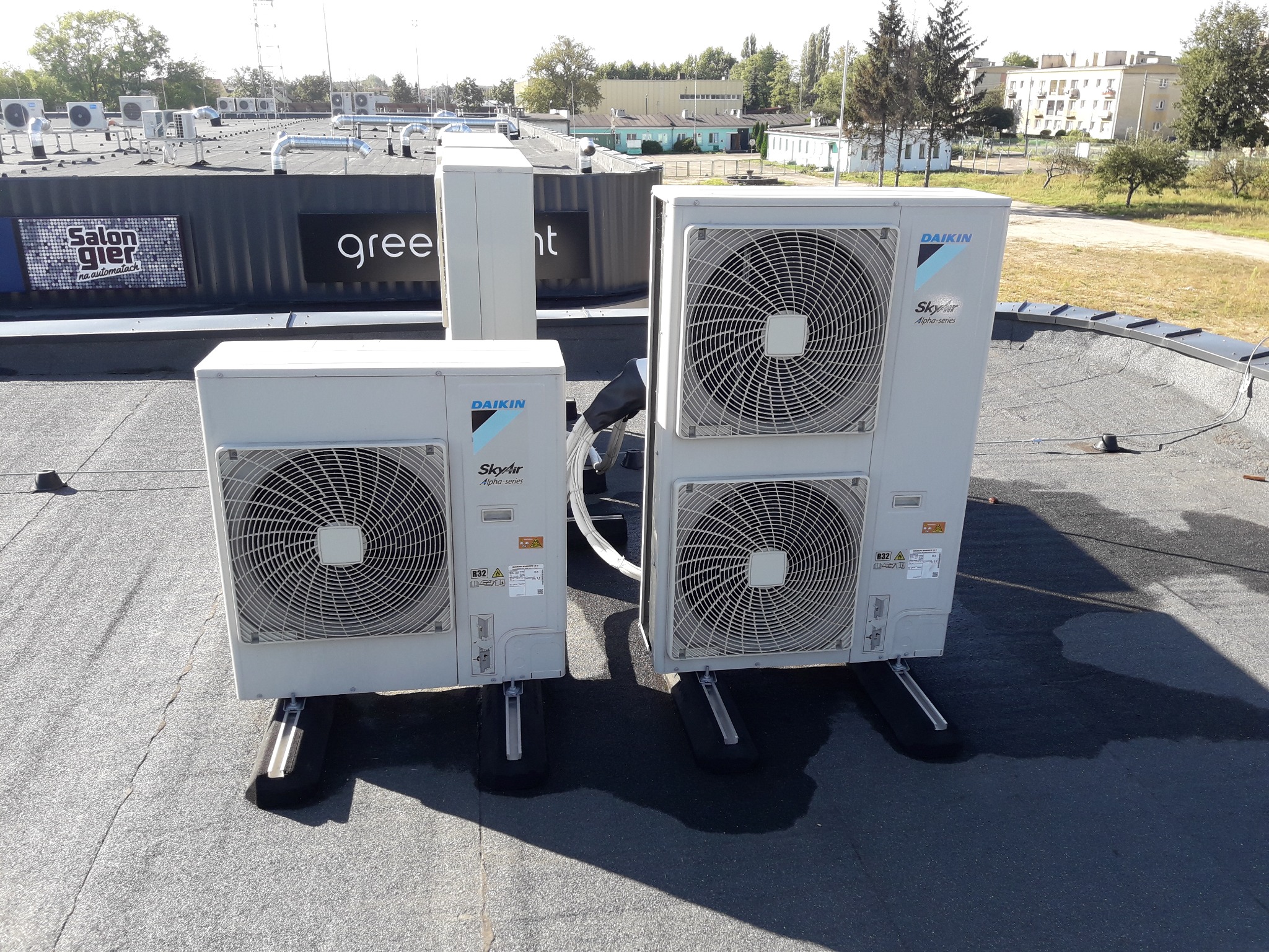 Dwa agregaty klimatyzacyjne Daikin SkyAir Alpha-series zamontowane na płaskim dachu z widocznymi przewodami i wspornikami antywibracyjnymi, w tle inne urządzenia i budynki.