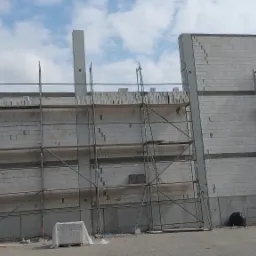 Wznoszenie ściany z bloczków na budowie, widoczne rusztowania i betonowe słupy konstrukcyjne, częściowo murowana ściana z widocznymi spoinami. Na dole leży czarna torba i paleta z materiałem...