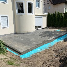 EKOINVEST 3 MIASTO SPÓŁKA Z OGRANICZONĄ ODPOWIEDZIALNOŚCIĄ - Wylewka betonowa tarasu przy domu jednorodzinnym, zabezpieczona niebieską folią hydroizolacyjną. Widoczne okna i rolety zewnętrzne budynku.