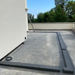 EKOINVEST 3 MIASTO SPÓŁKA Z OGRANICZONĄ ODPOWIEDZIALNOŚCIĄ - Izolacja przeciwwodna dachu płaskiego w trakcie prac. Widoczne czarne pasy izolacyjne tworzące ramę, kabel elektryczny i drabina oparta o ścianę budynku.