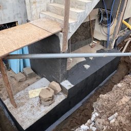 EKOINVEST 3 MIASTO SPÓŁKA Z OGRANICZONĄ ODPOWIEDZIALNOŚCIĄ - Fundament budynku w trakcie izolacji bitumicznej, widoczne schody betonowe, rury i wykop. Prace ziemne w toku, częściowo zabezpieczone deskami.