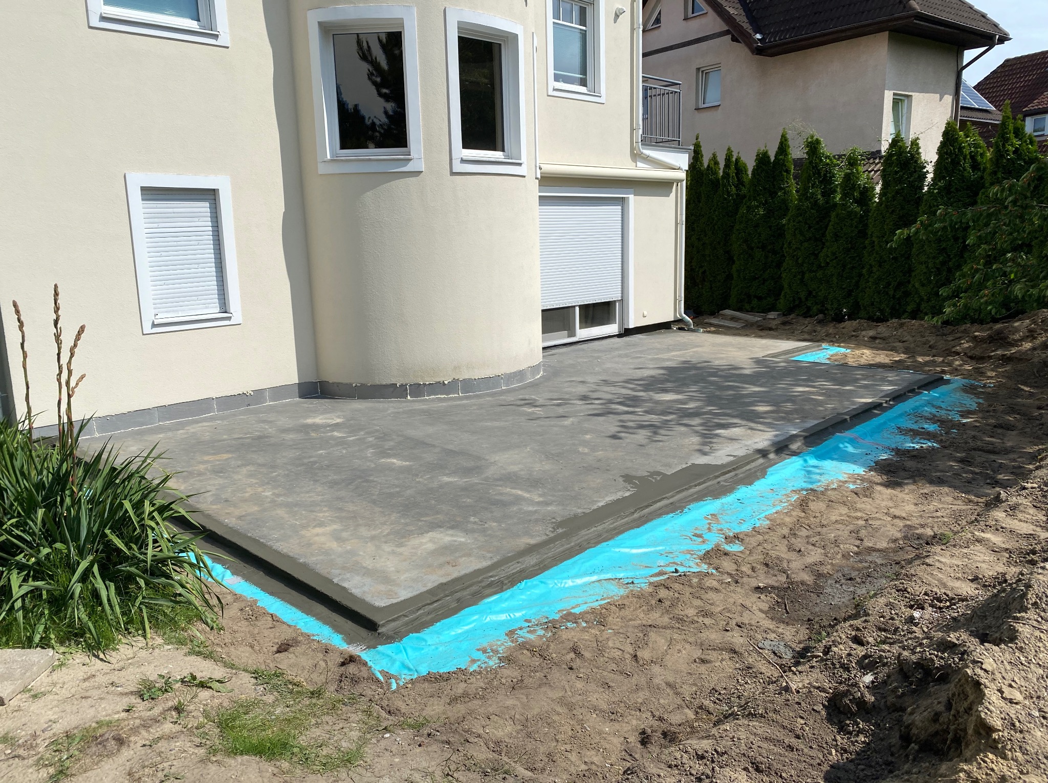 Wylewka betonowa tarasu przy domu jednorodzinnym, zabezpieczona niebieską folią hydroizolacyjną. Widoczne okna i rolety zewnętrzne budynku.