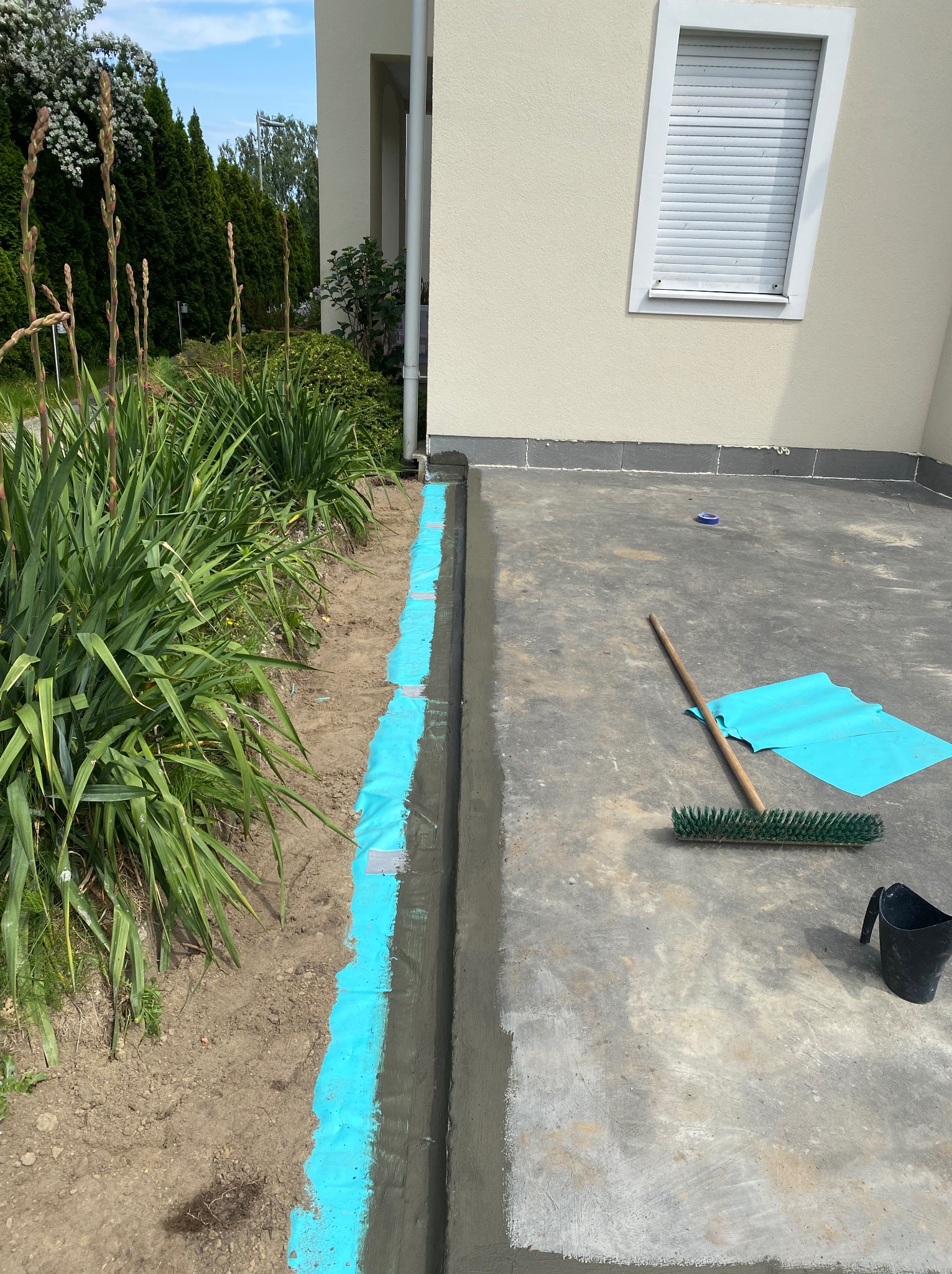 Etap izolacji fundamentów budynku jednorodzinnego z użyciem niebieskiej folii ochronnej i narzędzi, widok z góry, w tle zieleń i fragment elewacji.