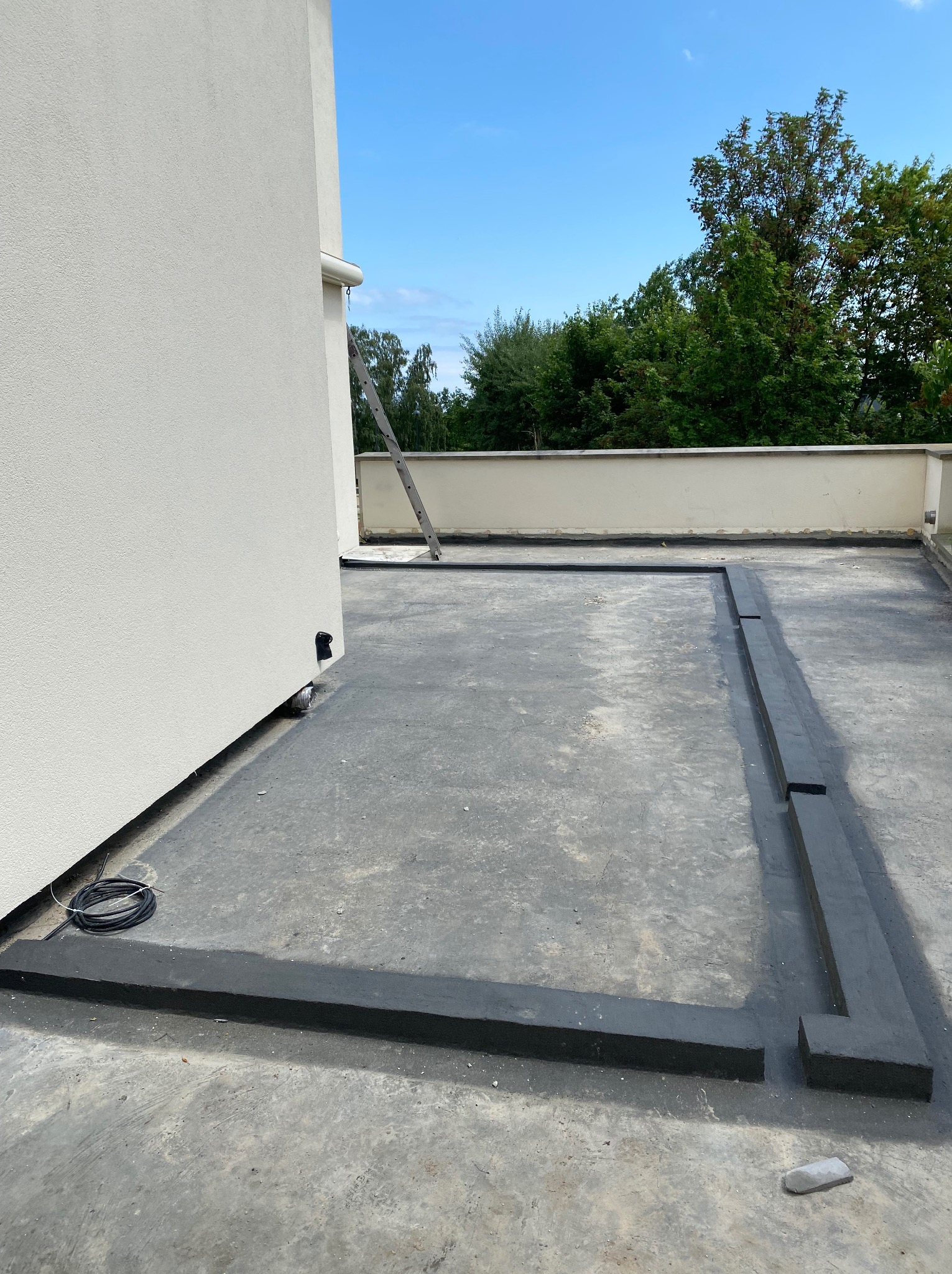 Izolacja przeciwwodna dachu płaskiego w trakcie prac. Widoczne czarne pasy izolacyjne tworzące ramę, kabel elektryczny i drabina oparta o ścianę budynku.