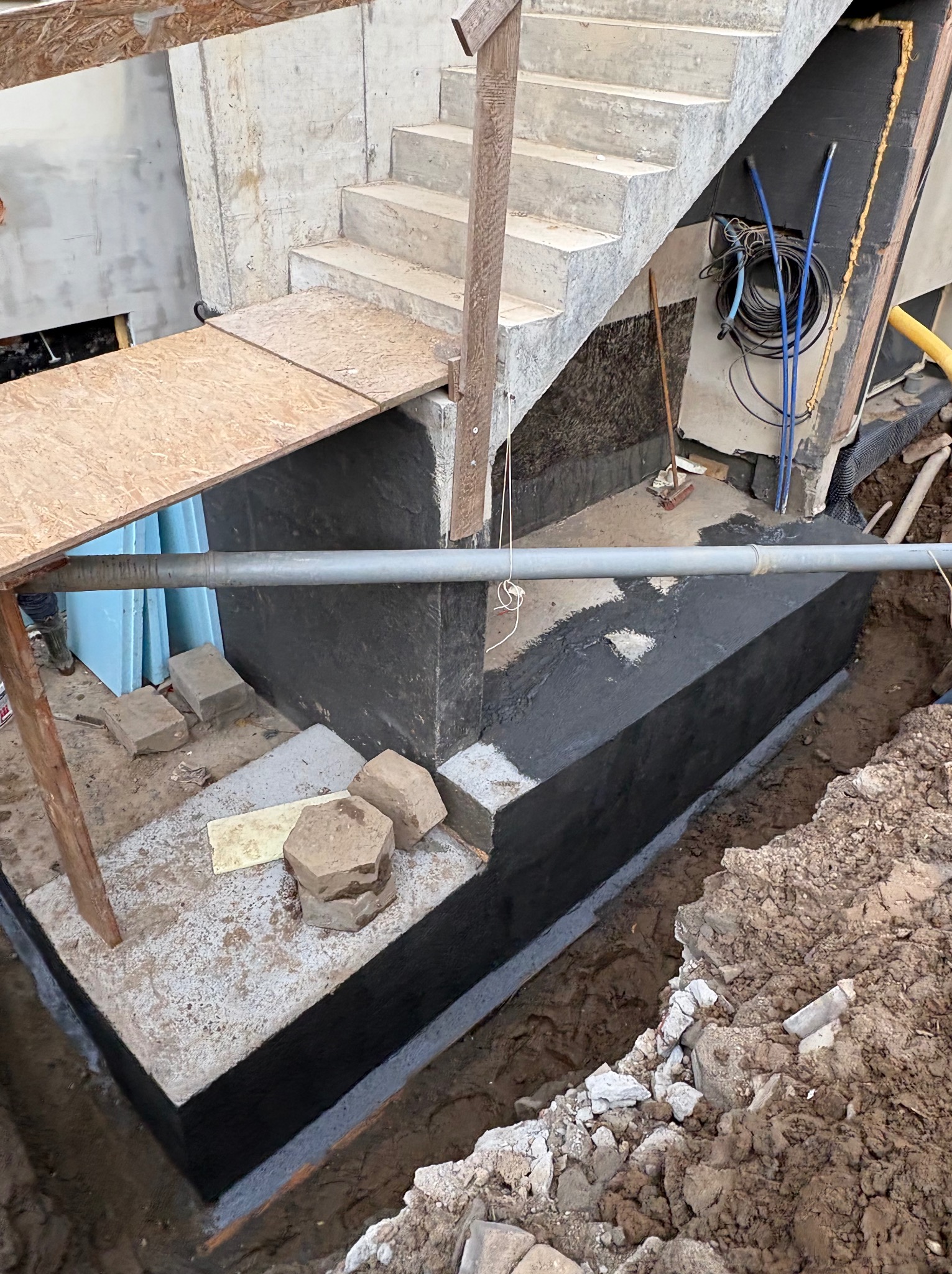 Fundament budynku w trakcie izolacji bitumicznej, widoczne schody betonowe, rury i wykop. Prace ziemne w toku, częściowo zabezpieczone deskami.