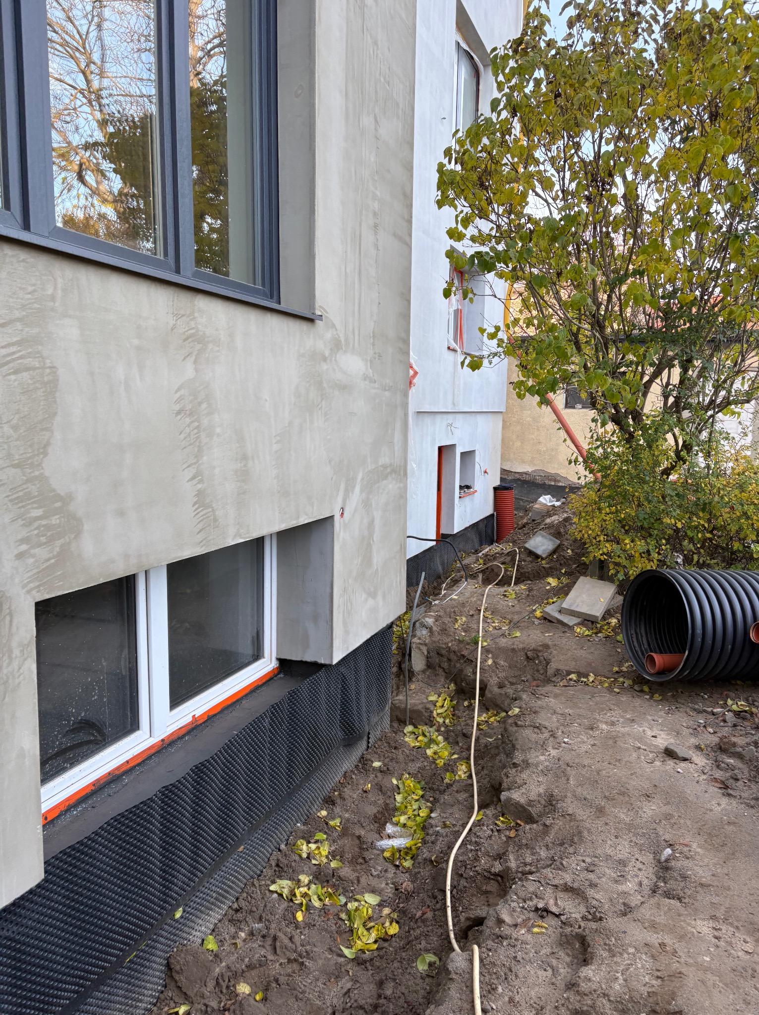 Częściowo ocieplony fundament budynku z widoczną czarną folią izolacyjną, wykopem i rurami drenażowymi. Jesienne liście na ziemi.