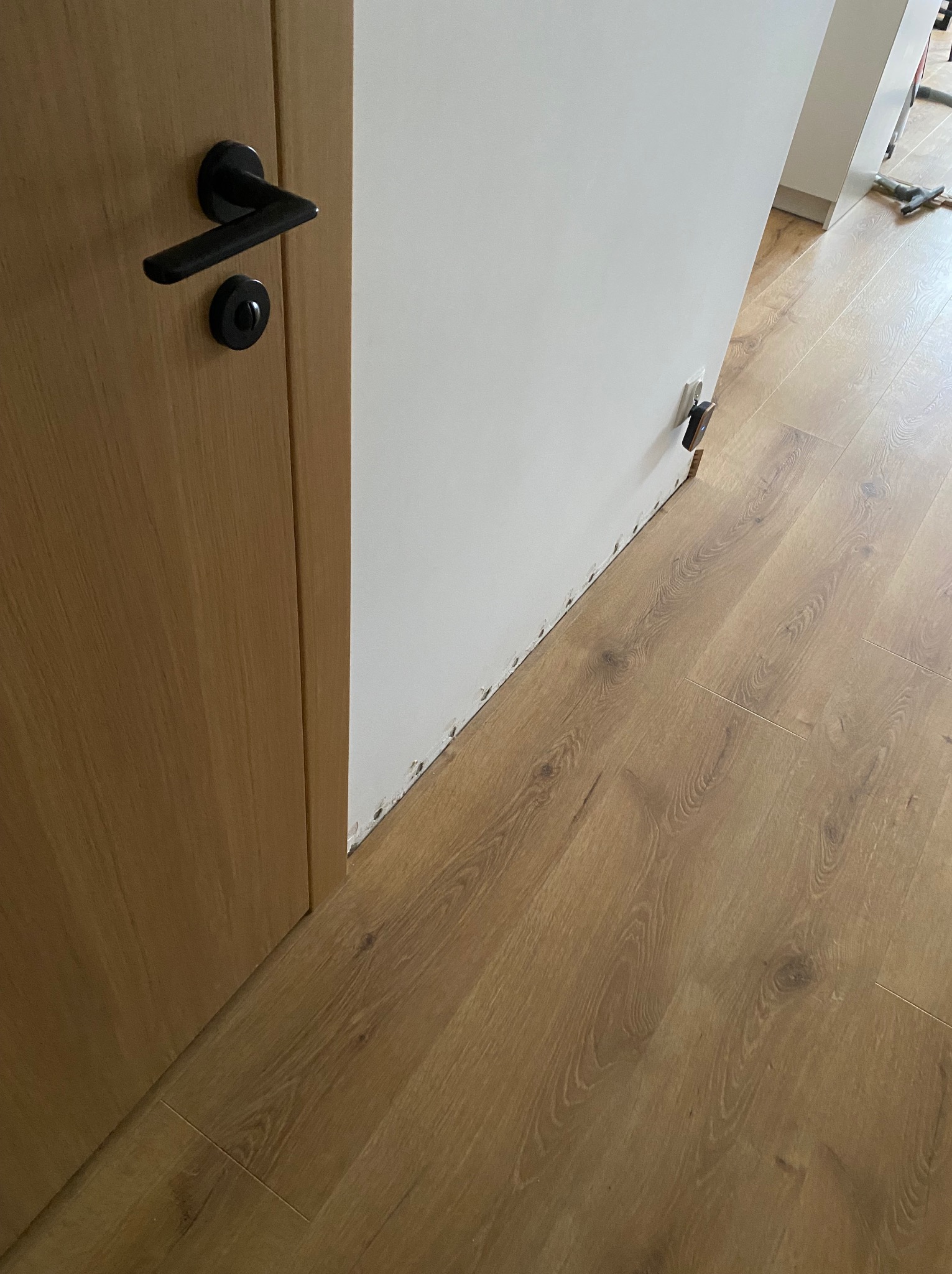 Montaż paneli podłogowych: widoczne łączenia, listwy przypodłogowe w trakcie mocowania, drewniane drzwi z czarną klamką. Jasne wnętrze w trakcie wykończenia.