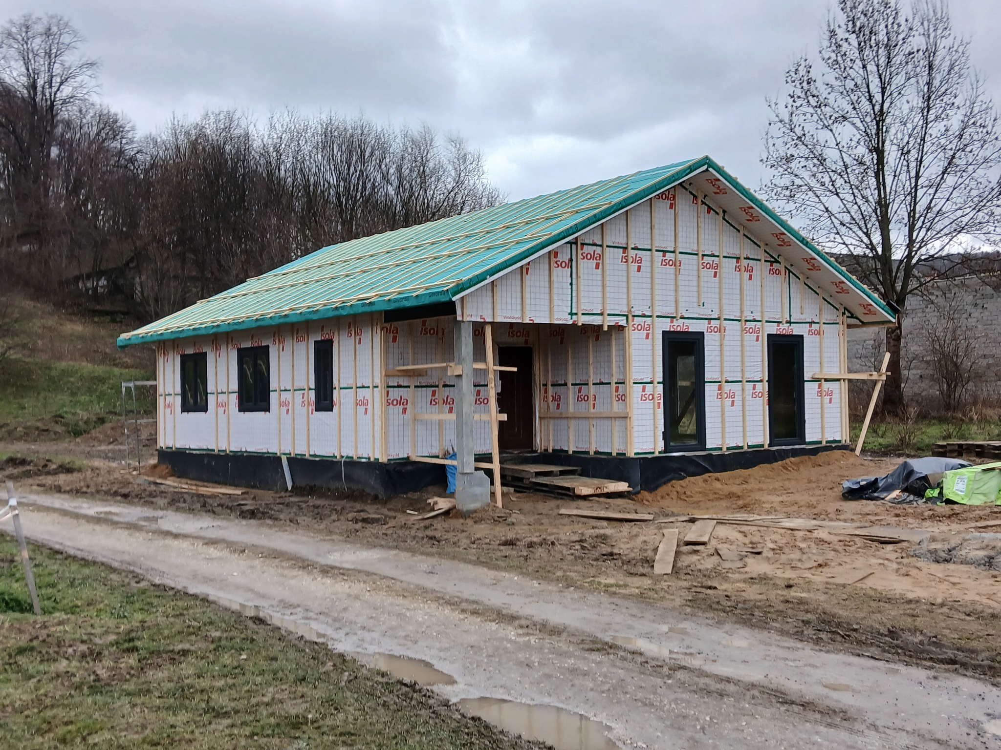 Budowa domu w technologii szkieletowej na etapie montażu izolacji i konstrukcji dachu, widoczne czarne okna, teren budowy z błotnistą drogą dojazdową.