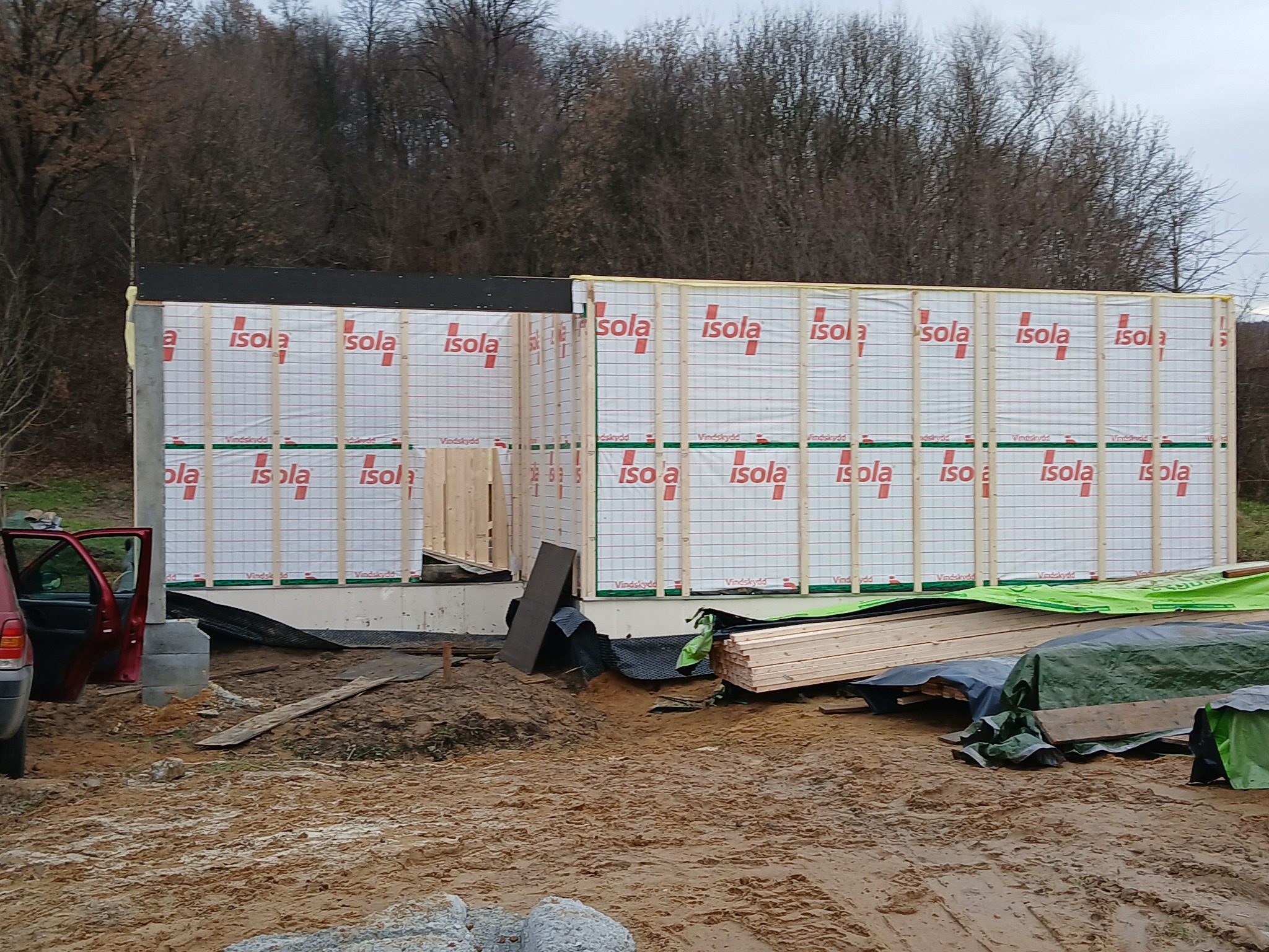 Wznoszenie surowej konstrukcji domu, widoczne ściany z izolacją i drewnianym szkieletem, częściowo otwarte, z widocznymi belkami, na zewnątrz rozłożone materiały budowlane, teren budowy z błotem...