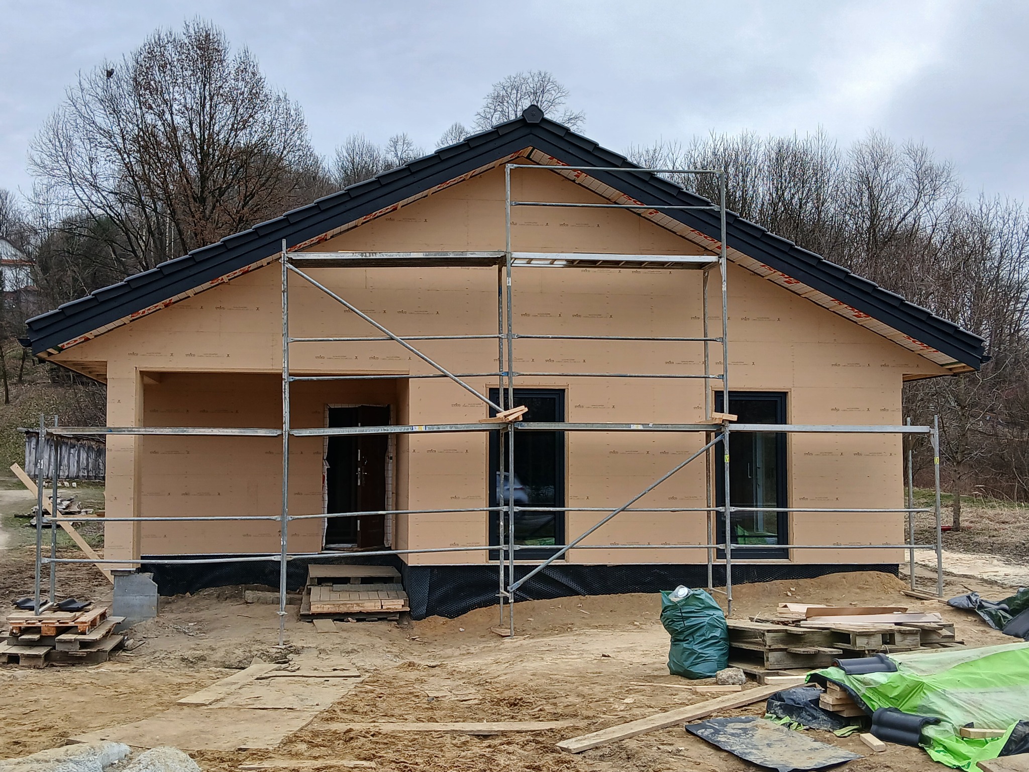 Budowa domu szkieletowego na etapie montażu izolacji zewnętrznej i rusztowań, widoczne czarne okna i dachówka, teren budowy w trakcie prac.