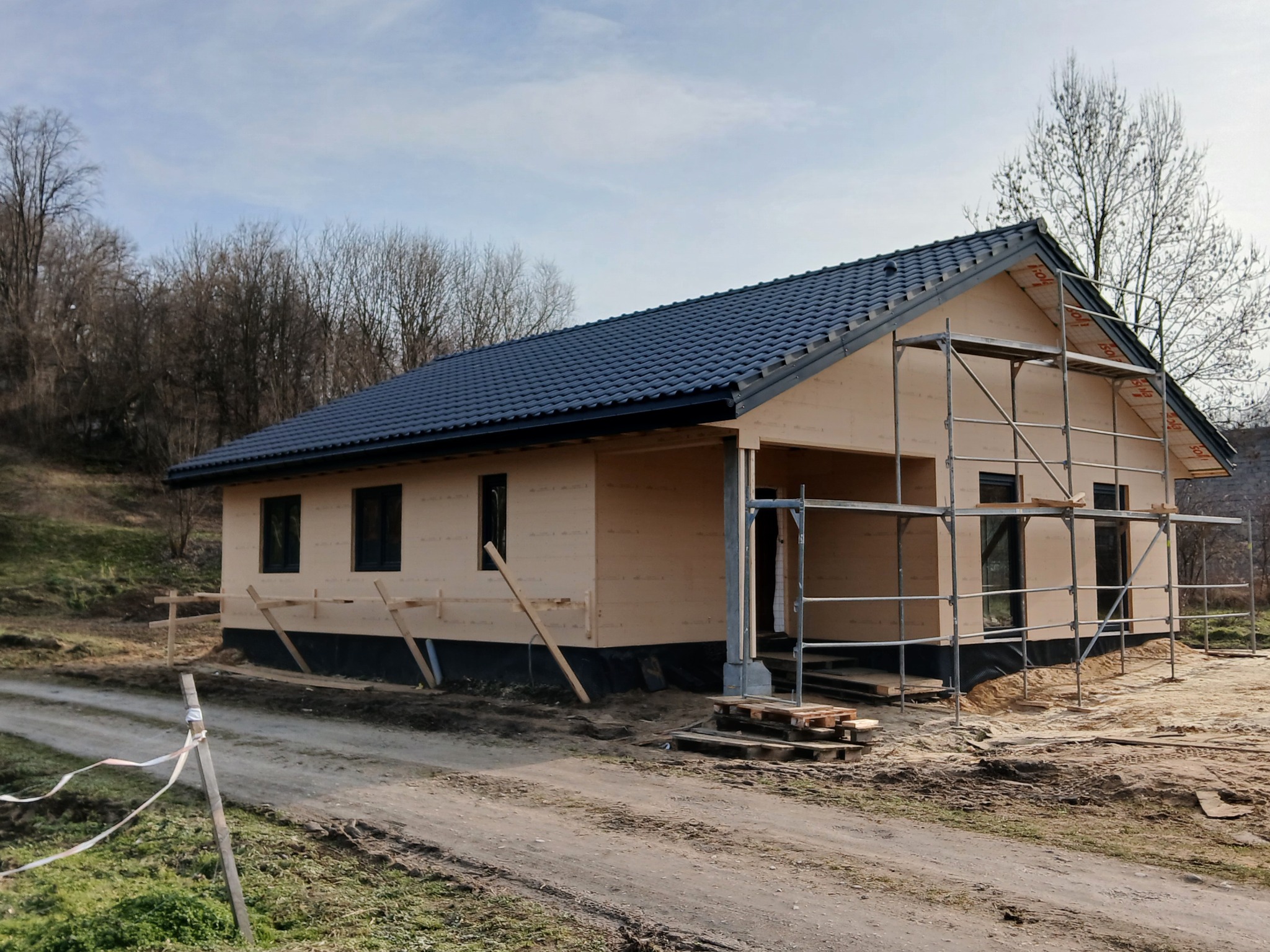 Surowy stan budynku z ciemną dachówką, ocieplonego jasną wełną mineralną, z rusztowaniem w tle i zabezpieczeniami przeciwwilgociowymi u dołu, na tle drzew i nieba.