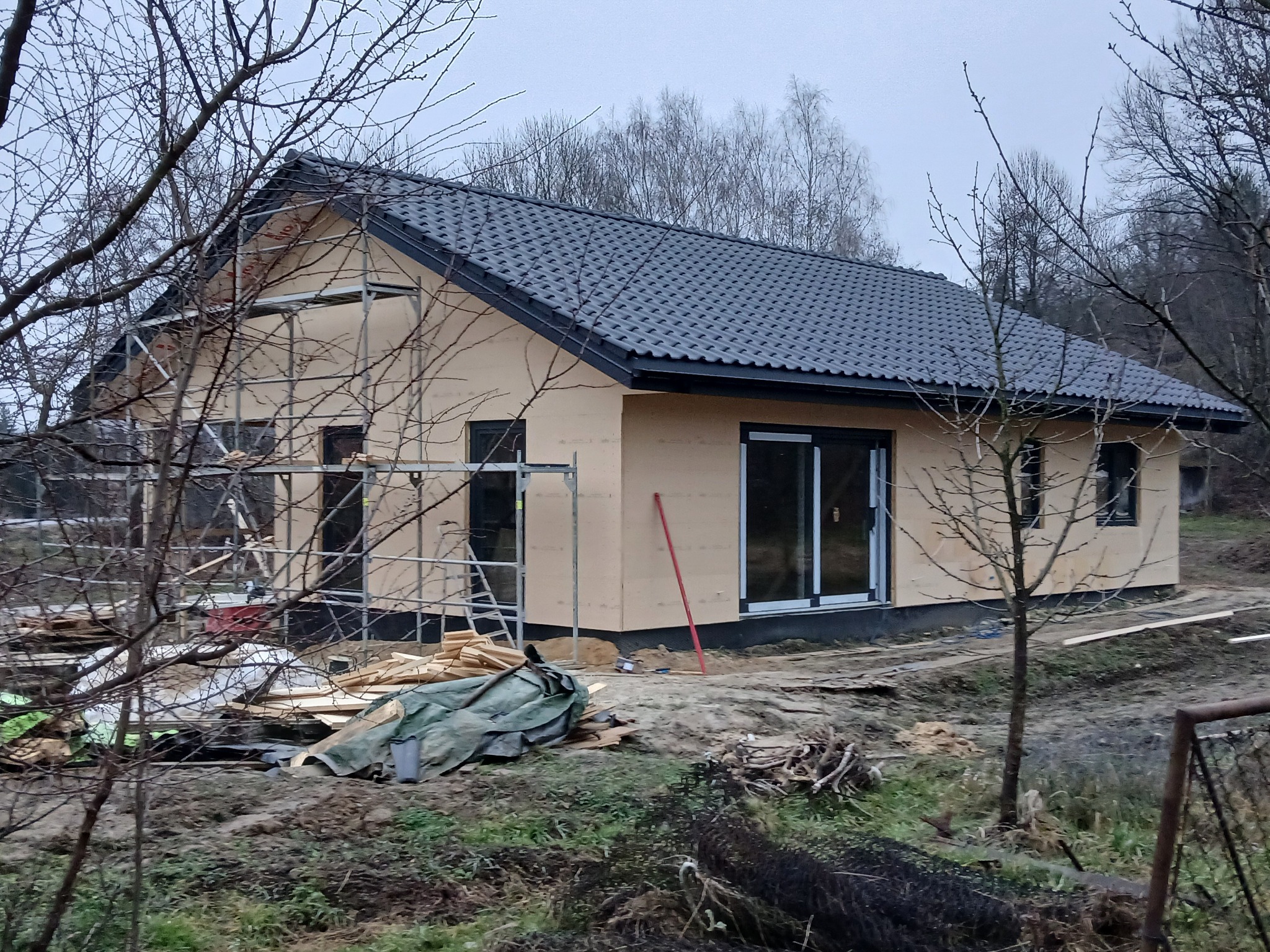 Budowa domu w technologii szkieletowej z widocznym ociepleniem ścian zewnętrznych, ciemna dachówka, stolarka okienna w kolorze czarnym, teren budowy z materiałami budowlanymi i rusztowaniami.