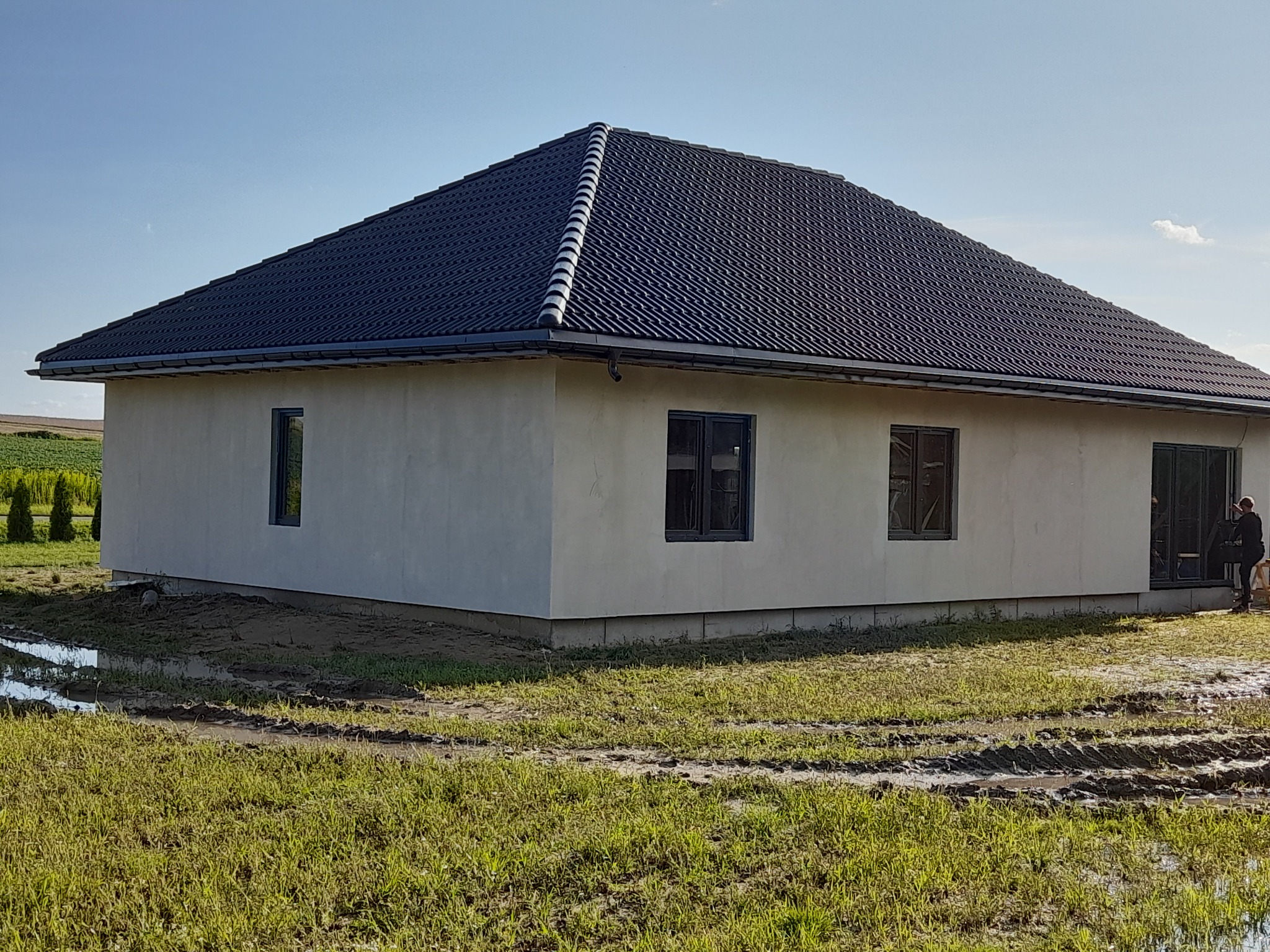 Niedawno wybudowany parterowy budynek z ciemną dachówką, jasnymi ścianami i ciemnymi ramami okiennymi, otoczony trawnikiem z błotnistymi śladami opon.