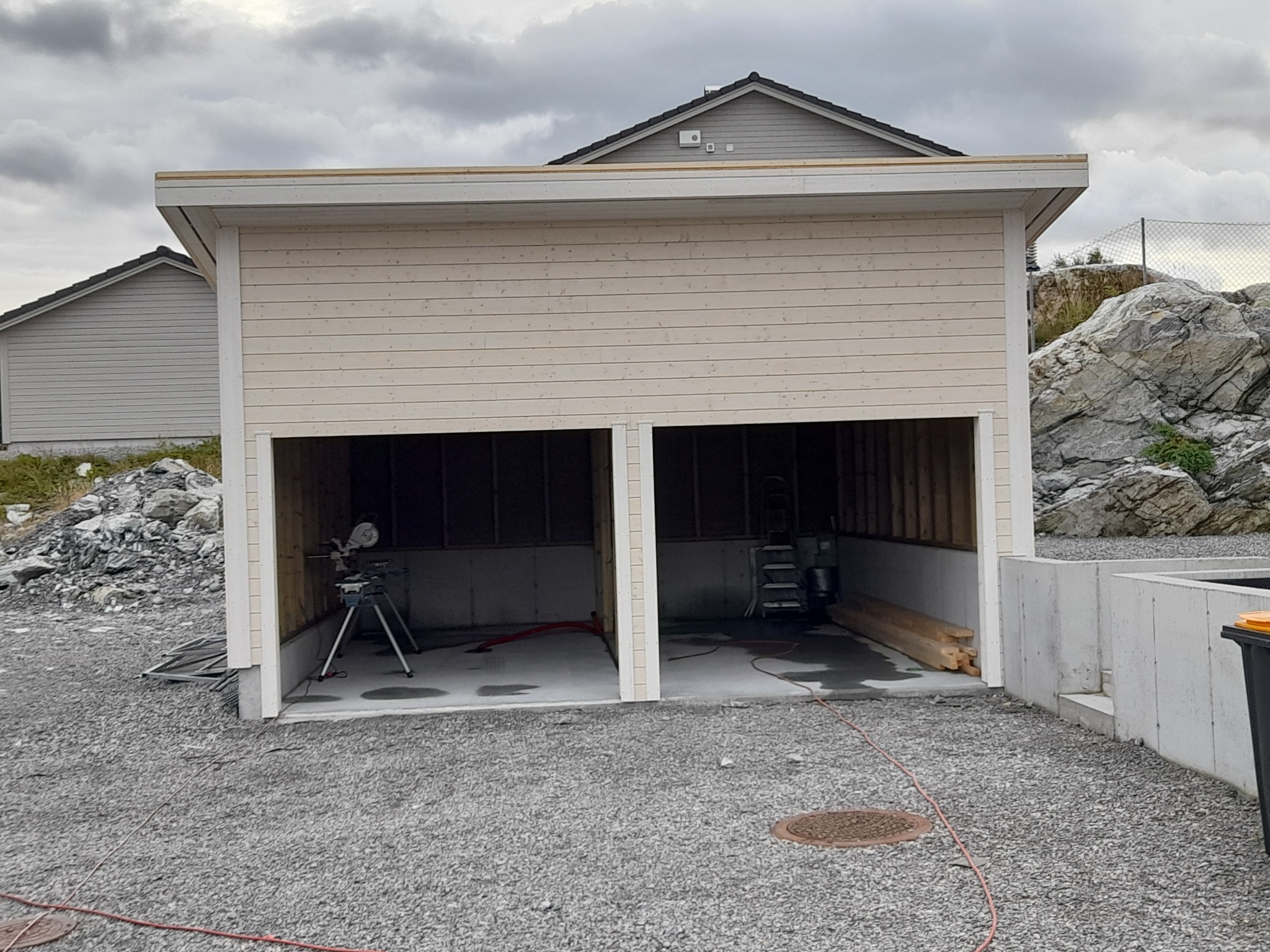 Wznoszony garaż dwustanowiskowy z jasnego drewna, widoczne narzędzia i materiały budowlane wewnątrz, teren wokół wysypany żwirem.