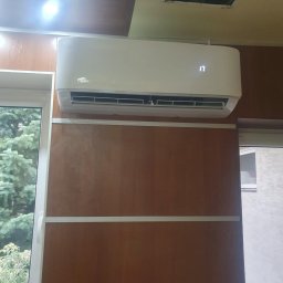 MG-Klima TABAKA Marek Giemza - Biała klimatyzacja marki Vivax zamontowana na ścianie z boazerii, obok okna z widokiem na zieleń.