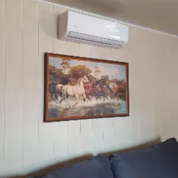 Biały klimatyzator marki Rotenso zamontowany nad obrazem z motywem koni, na ścianie wyłożonej panelami.