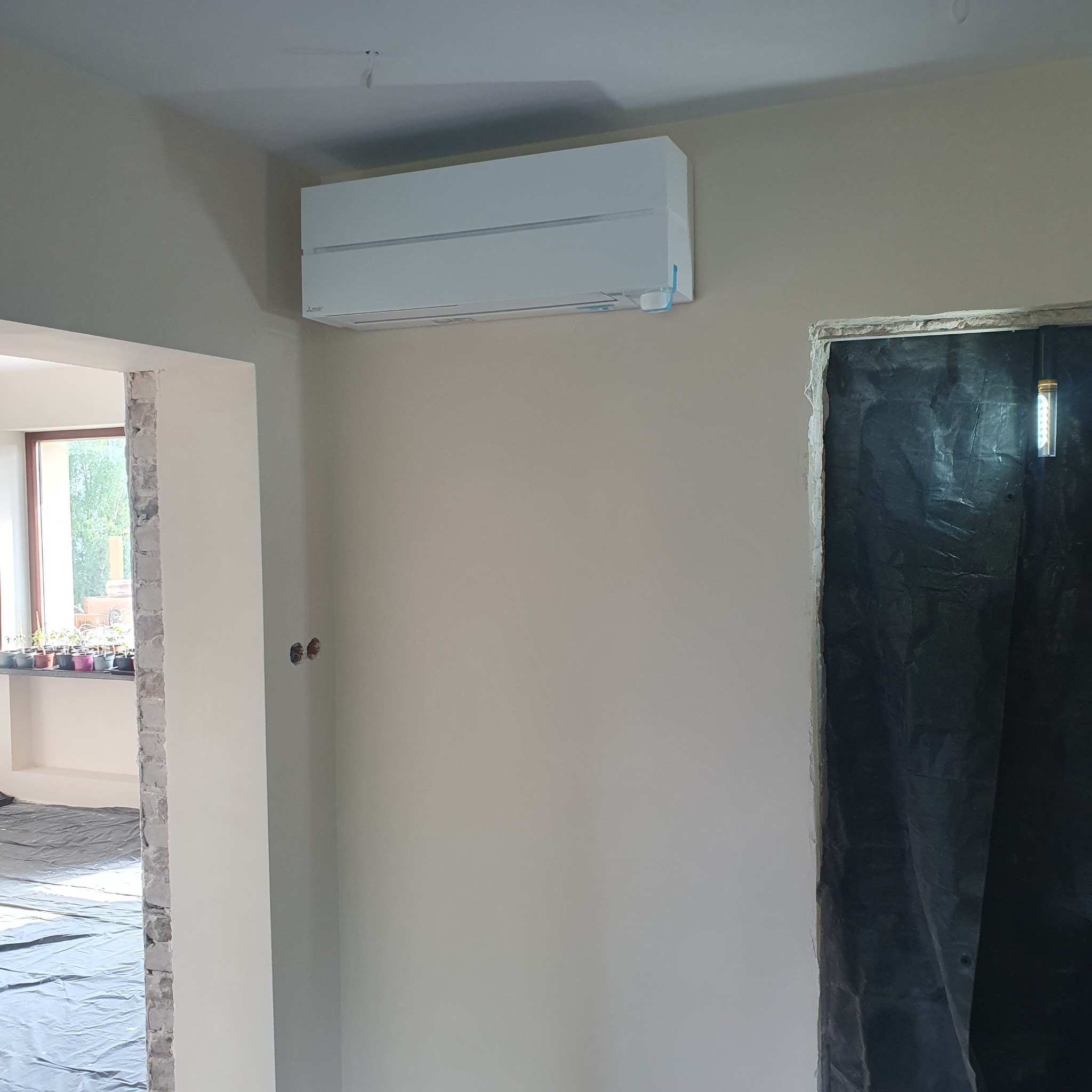 Biała jednostka klimatyzacji Mitsubishi Electric zamontowana na ścianie w remontowanym pokoju, widoczne przygotowane otwory instalacyjne, okno z roślinami w tle i zabezpieczone folią drzwi.