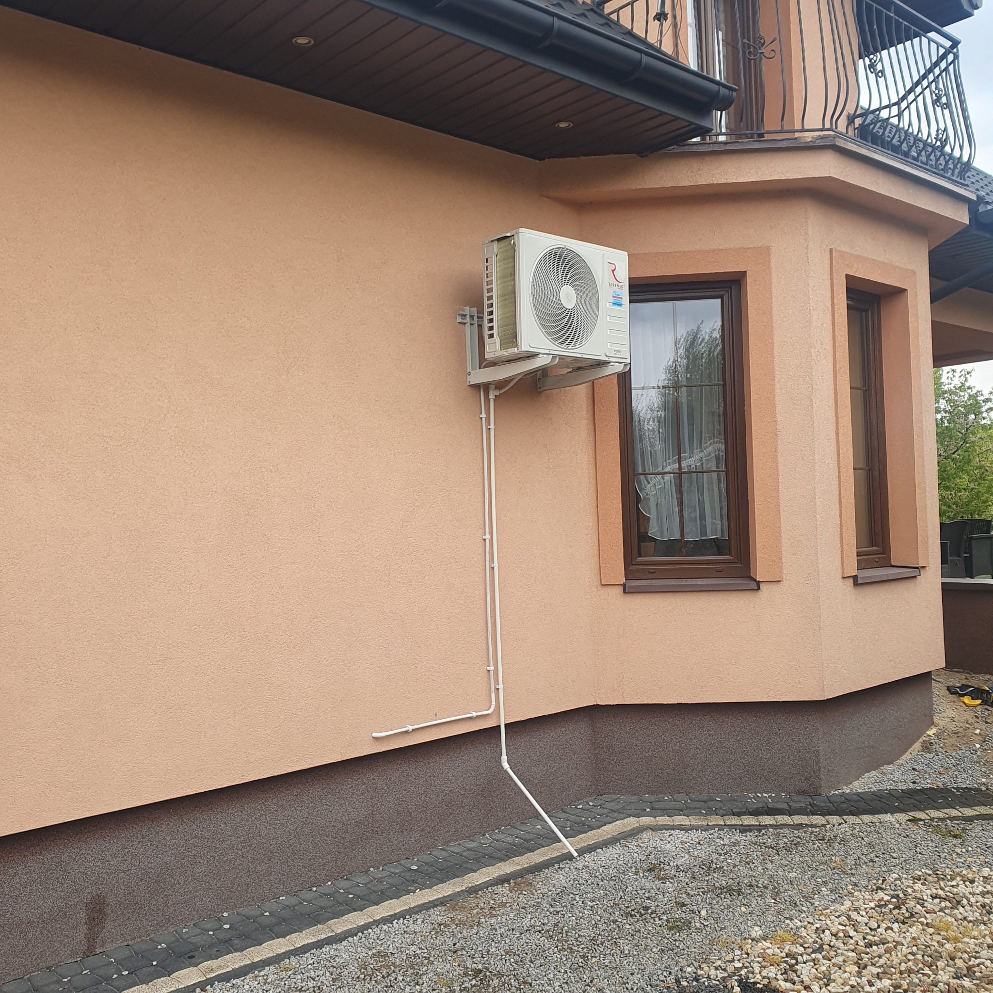 Biała jednostka klimatyzacji zewnętrznej zamontowana na beżowej ścianie domu, widoczne rury odprowadzające skropliny i okno z brązową ramą.