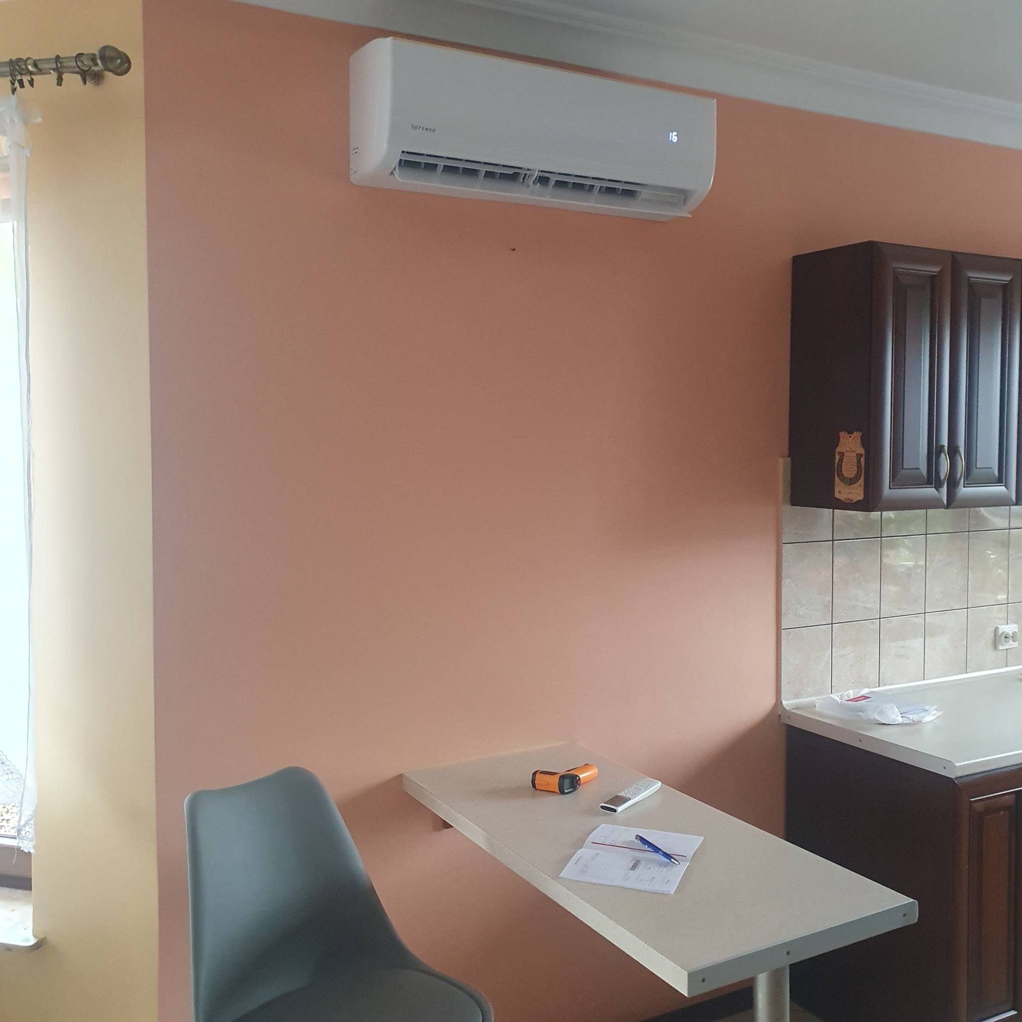 Biały klimatyzator Rotenso zamontowany na pomarańczowej ścianie w kuchni, widoczny termometr laserowy, pilot i dokumenty na składanym blacie.