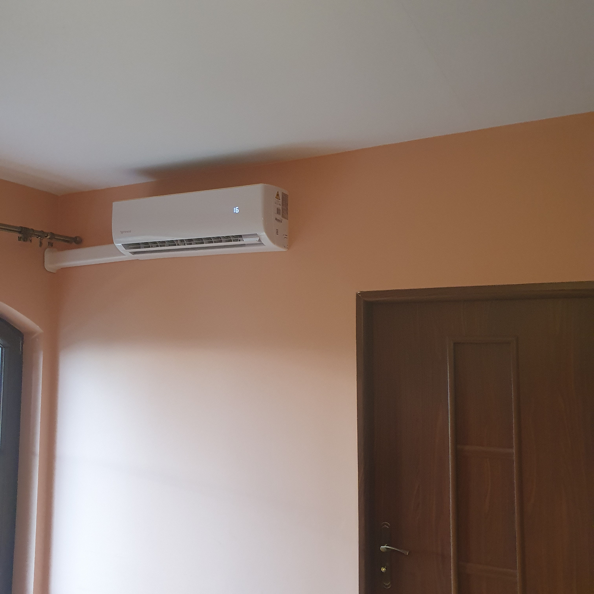 Biała jednostka klimatyzacyjna z wyświetlaczem temperatury (16 stopni) zamontowana na pomarańczowej ścianie w pomieszczeniu z brązowymi drzwiami.