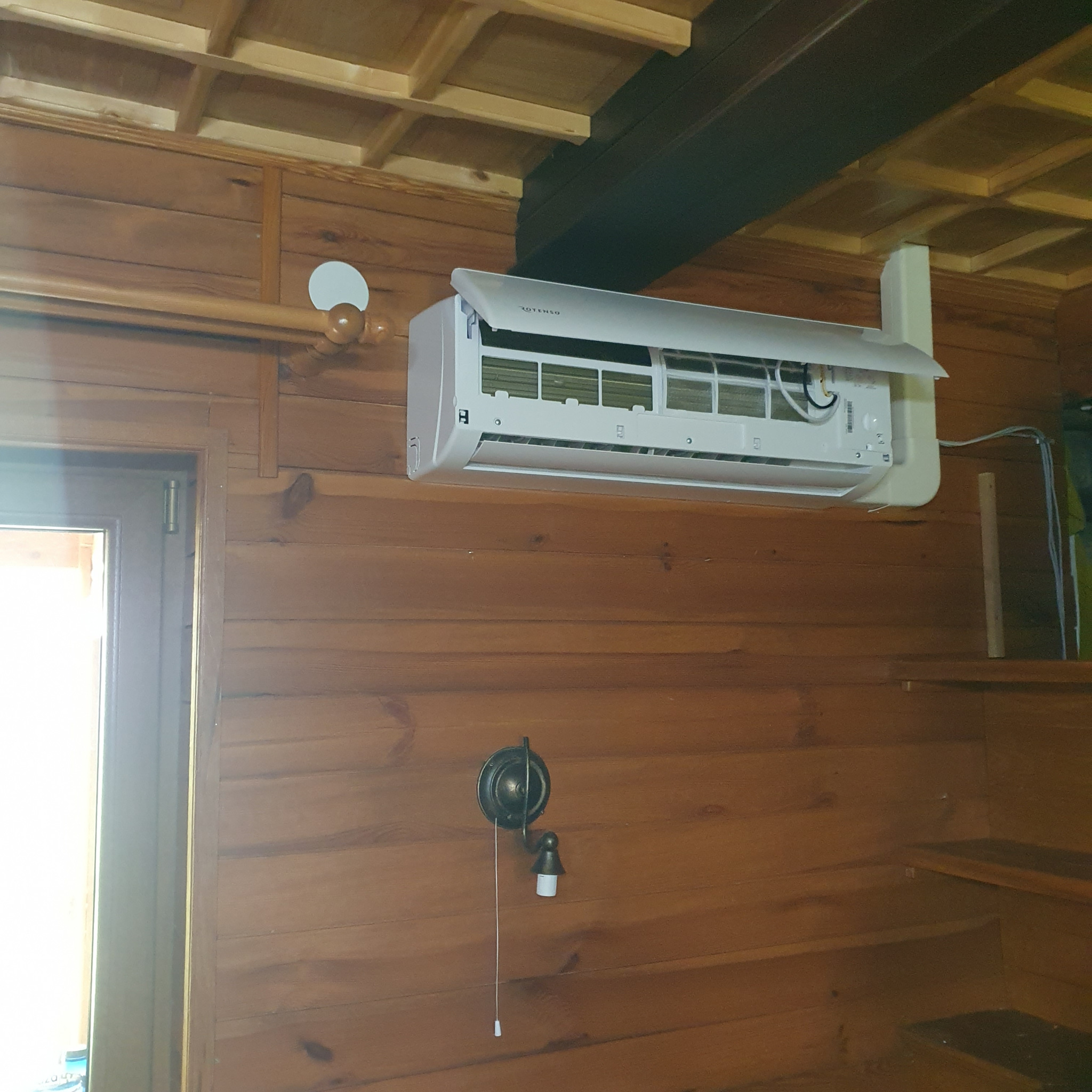 Biała jednostka klimatyzacyjna marki Rotenso zamontowana na drewnianej ścianie wewnątrz pomieszczenia, widoczne filtry i przewody, obok lampka ścienna z włącznikiem sznurkowym.