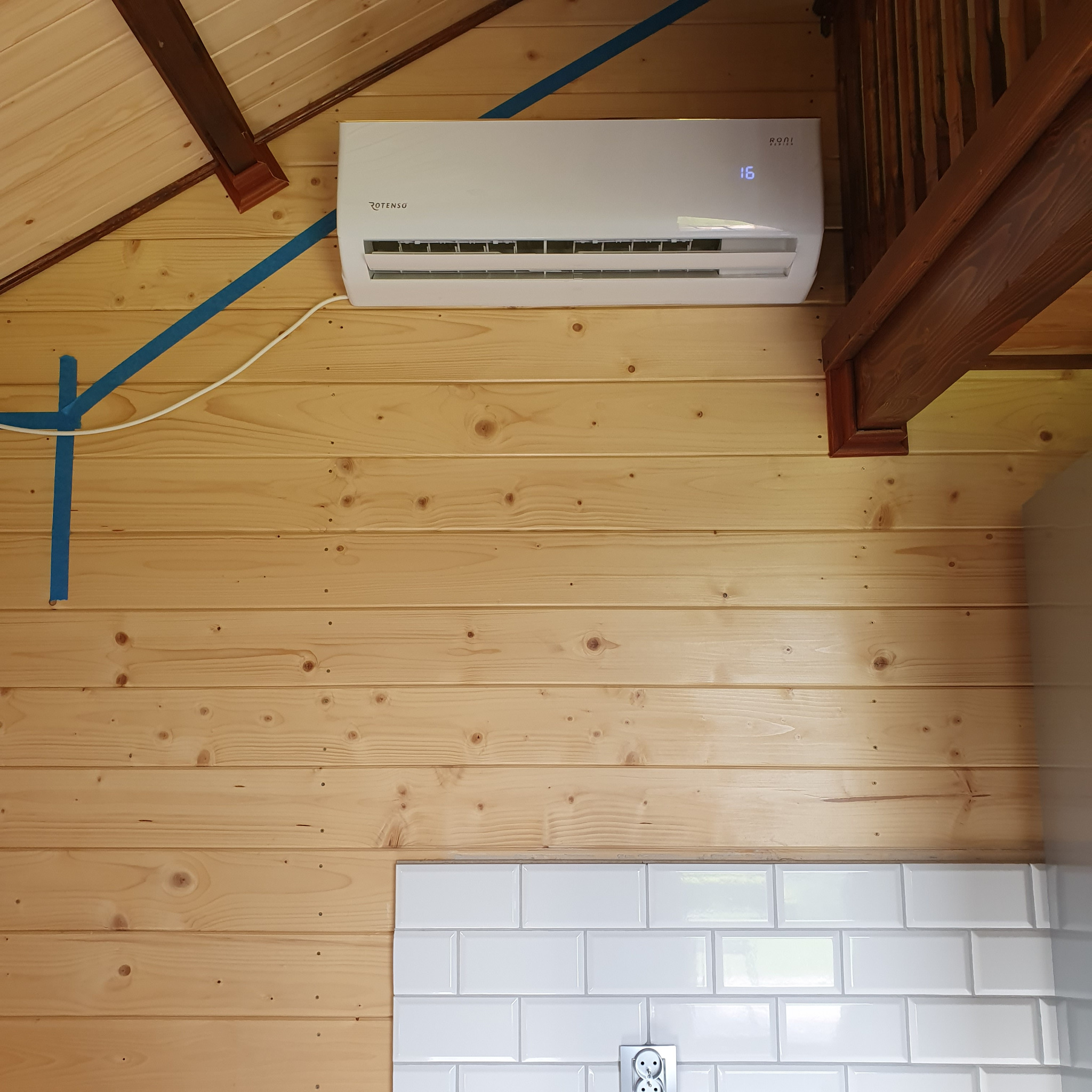 Biała jednostka klimatyzacyjna Rotenso zamontowana na drewnianej ścianie w domu, widoczna instalacja elektryczna zabezpieczona niebieską taśmą malarską, fragment schodów drewnianych i białe płytki...