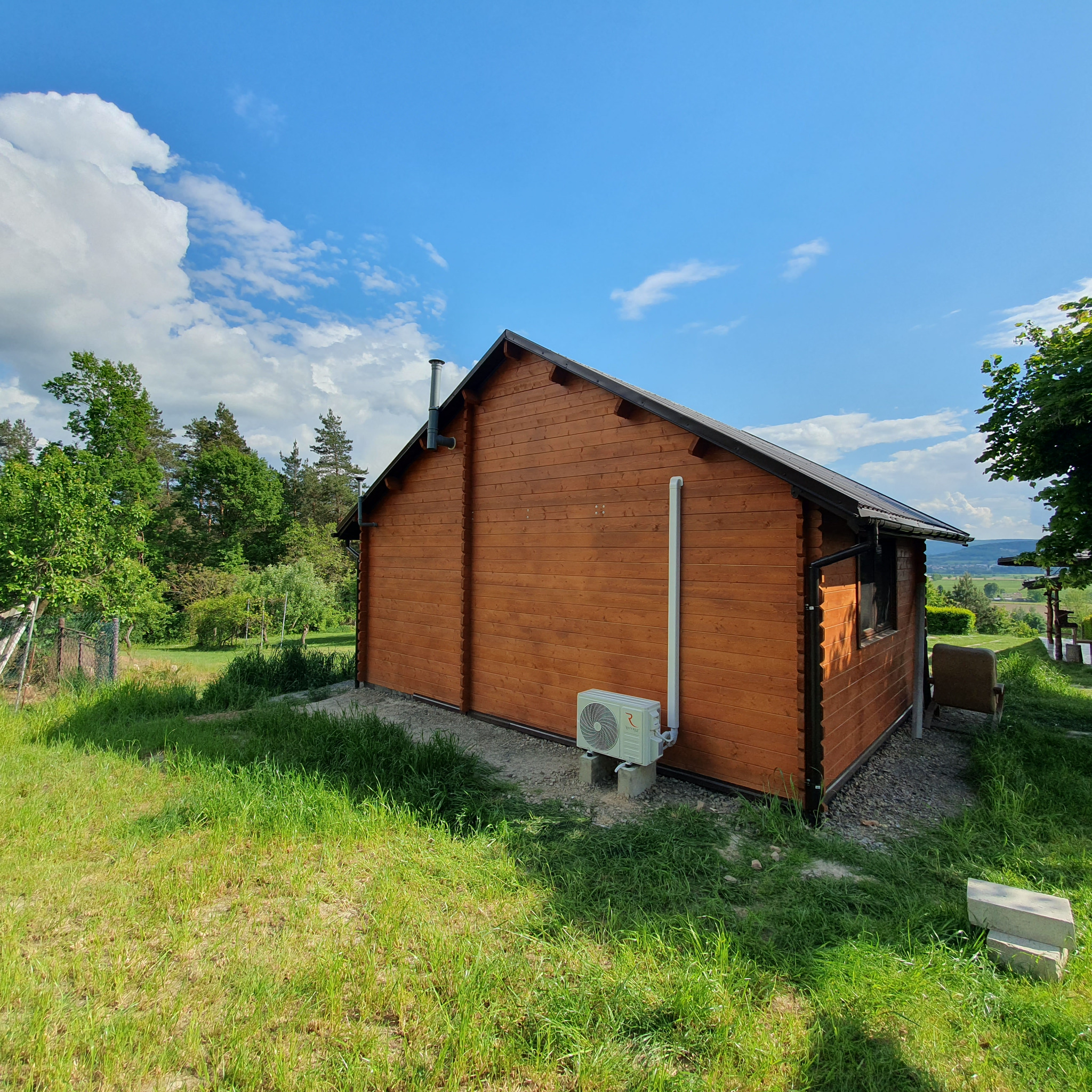 Drewniany domek z zamontowaną jednostką zewnętrzną klimatyzacji na betonowych bloczkach, widoczna rura odprowadzająca skropliny, na tle zielonego trawnika i błękitnego nieba.