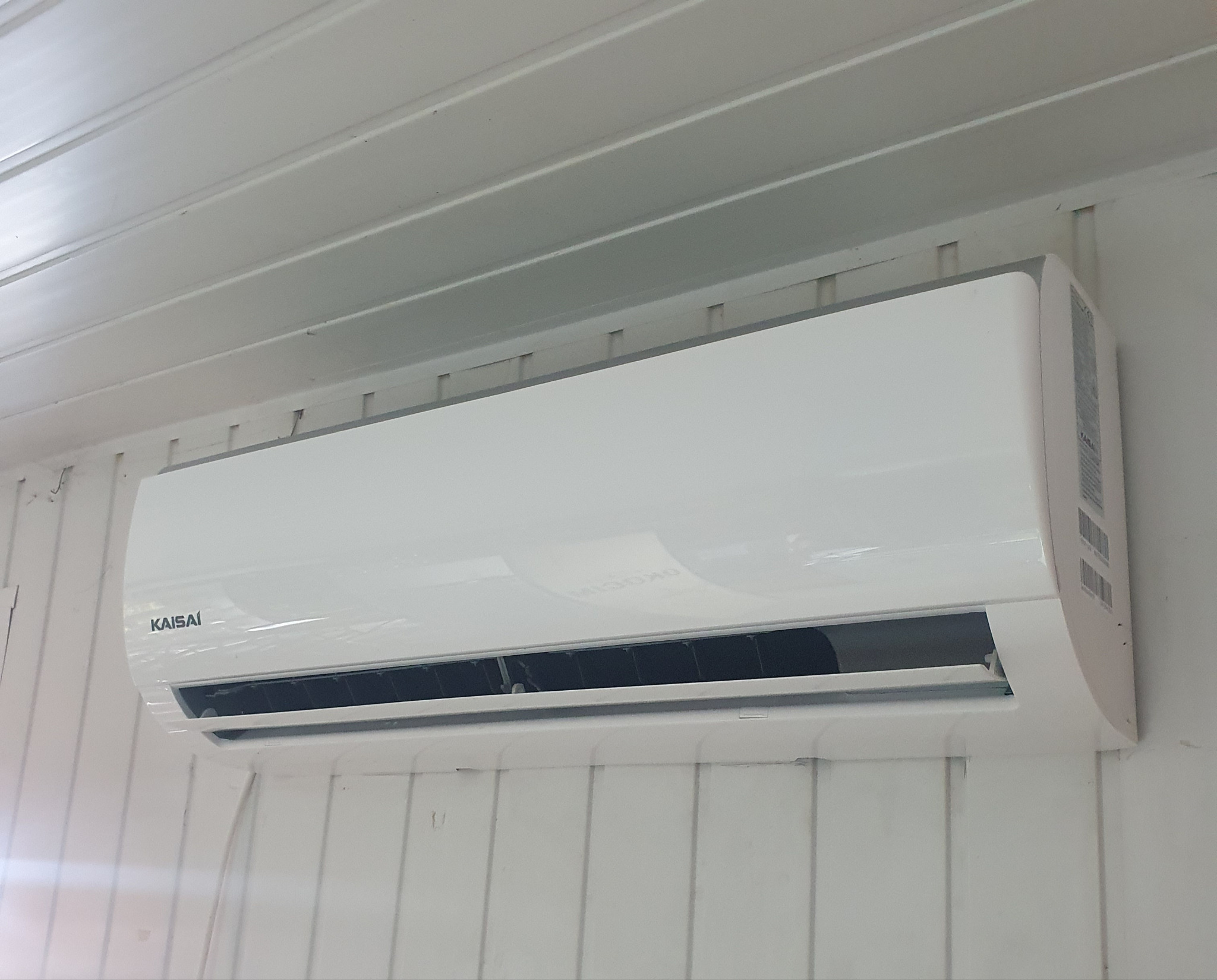 Biały klimatyzator Kaisai zamontowany na ścianie z białych paneli boazeryjnych.