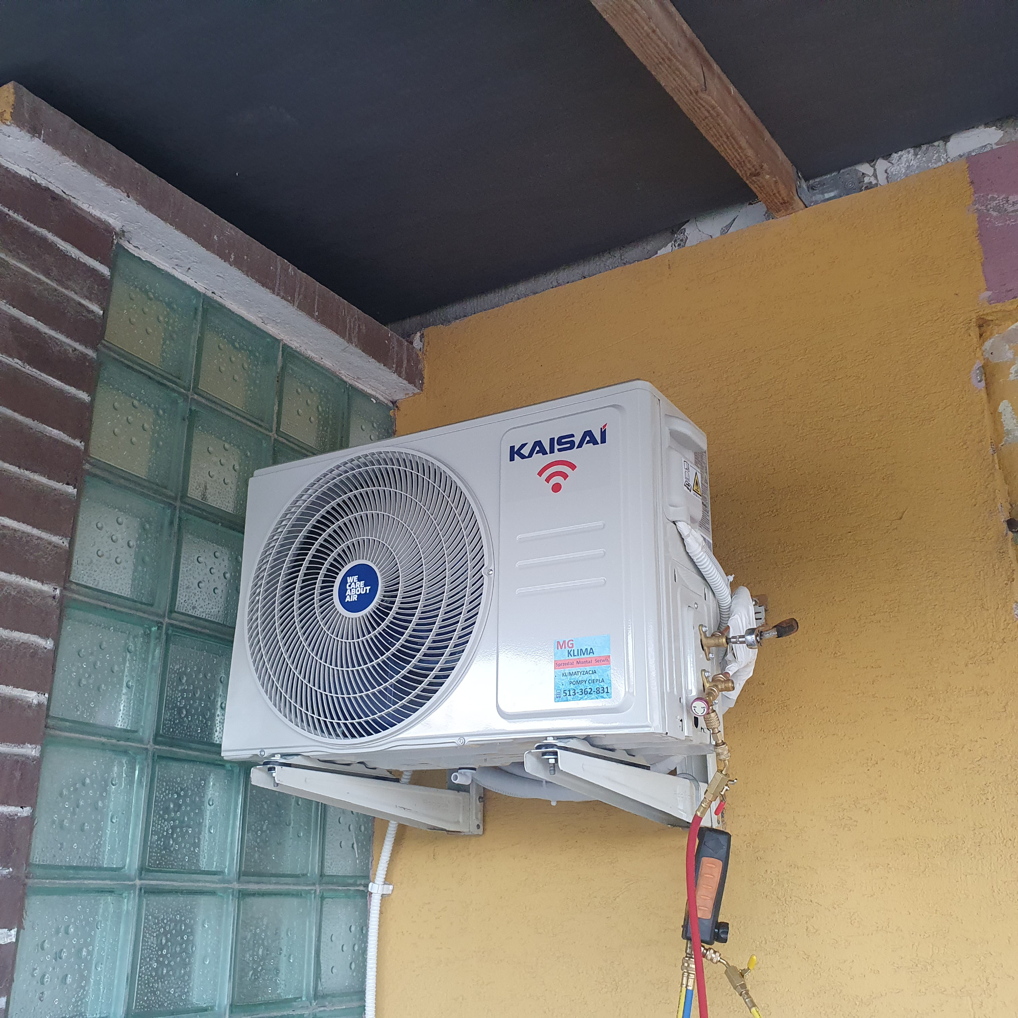 Zewnętrzna jednostka klimatyzacji Kaisai zamontowana na ścianie budynku w Ostrowcu Świętokrzyskim, z podłączonymi narzędziami serwisowymi do pomiaru ciśnienia czynnika chłodniczego.