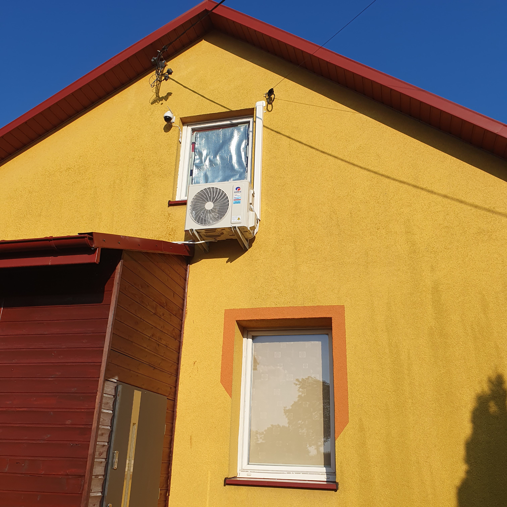 Żółty dom z zainstalowanym klimatyzatorem marki Gree pod oknem zabezpieczonym folią przeciwsłoneczną, widoczna kamera monitoringu i brązowe elementy wykończeniowe.