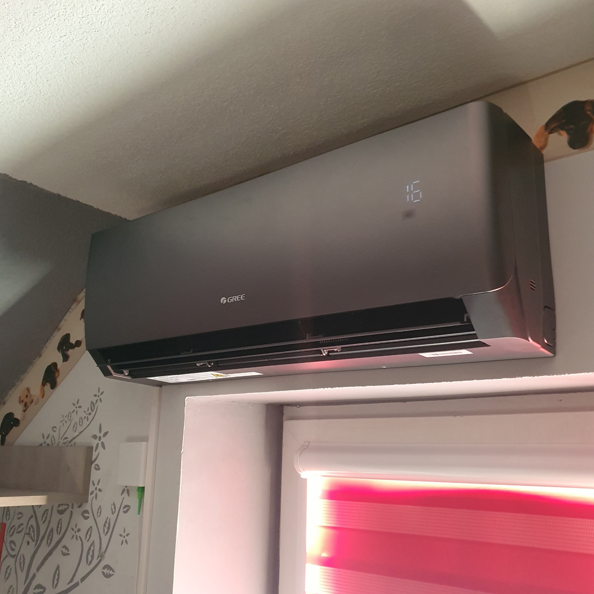 Szara jednostka klimatyzacji marki Gree zamontowana na ścianie w pomieszczeniu z dekoracjami ściennymi i roletami w oknie.