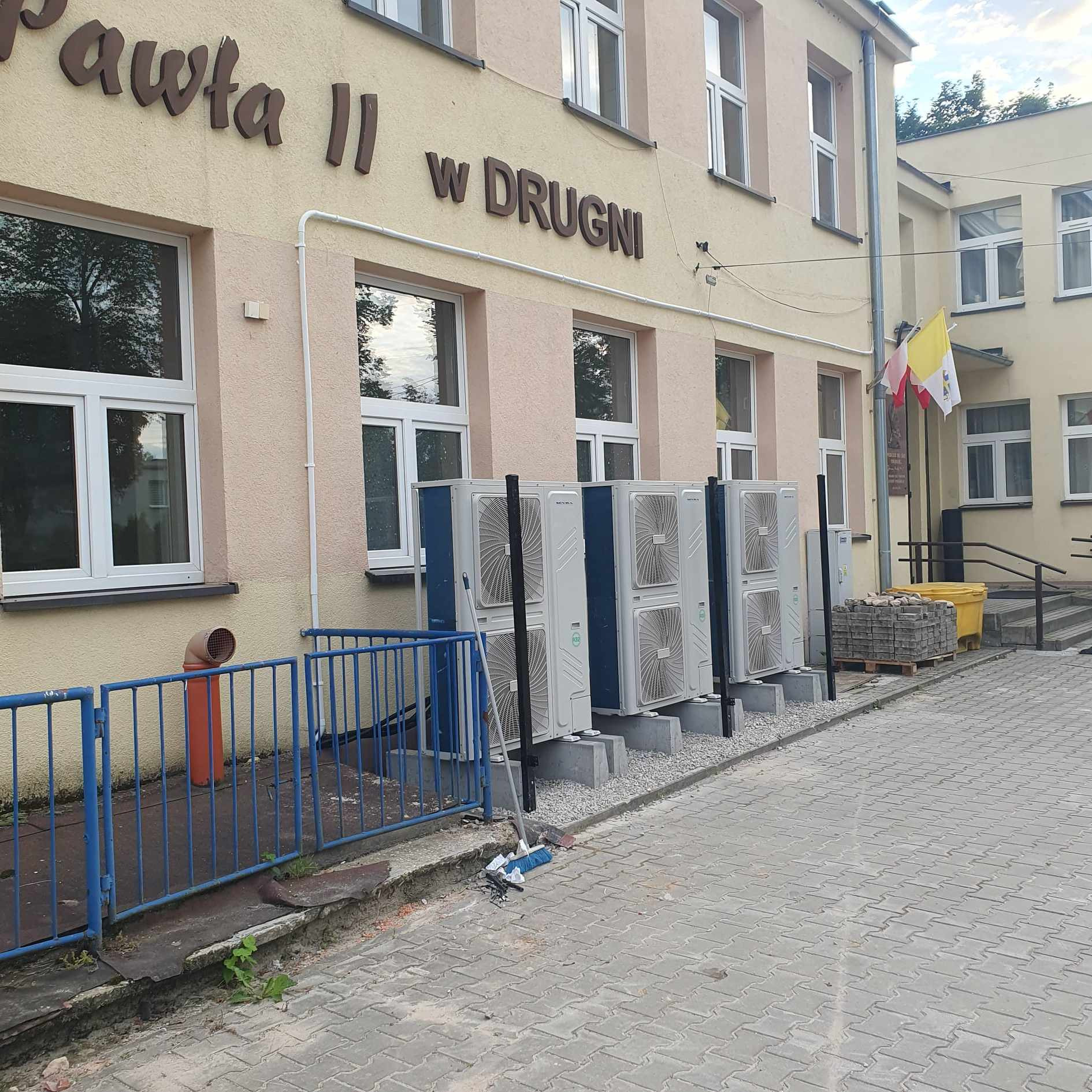 Zewnętrzne jednostki klimatyzacji zamontowane wzdłuż ściany budynku szkoły z napisem 'Pawła II w DRUGNI', widoczna flaga państwowa, obok leżą kostki brukowe i stoi miotła.