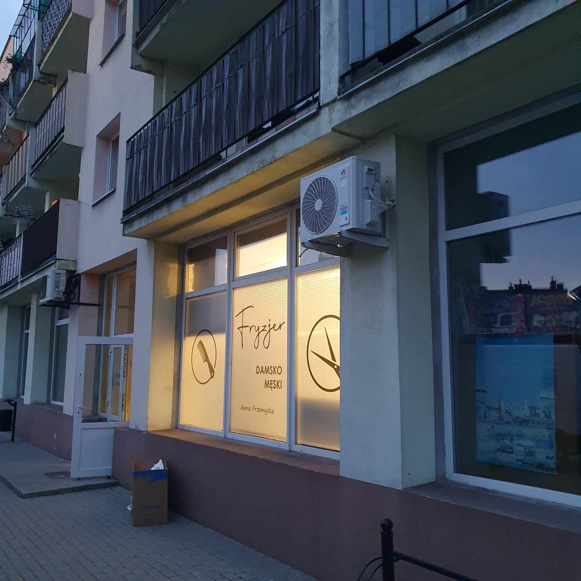 Zewnętrzny klimatyzator zamontowany nad oknem lokalu usługowego z napisem 'Fryzjer damsko-męski Anna Przemyska', grafiki grzebienia i nożyczek na oknie, widok z chodnika.