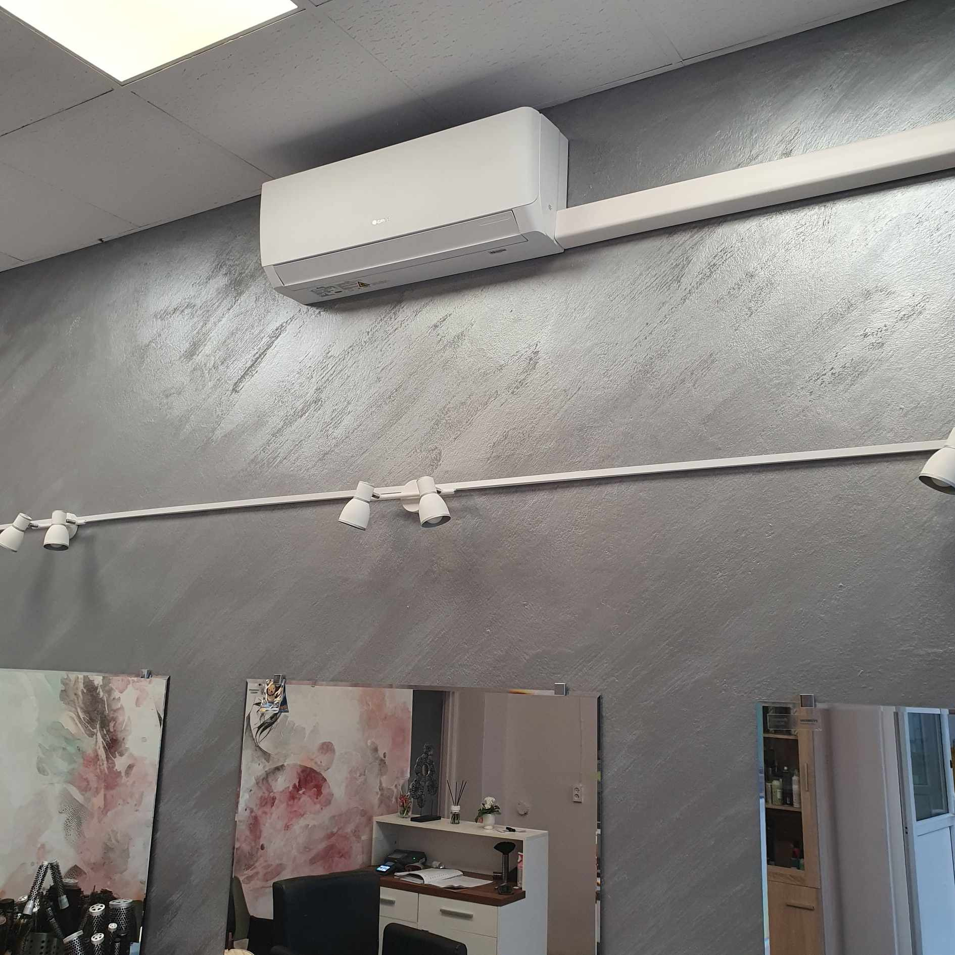 Biała jednostka wewnętrzna klimatyzacji zamontowana na szarej, teksturowanej ścianie w salonie fryzjerskim, widoczne oświetlenie szynowe i lustra.